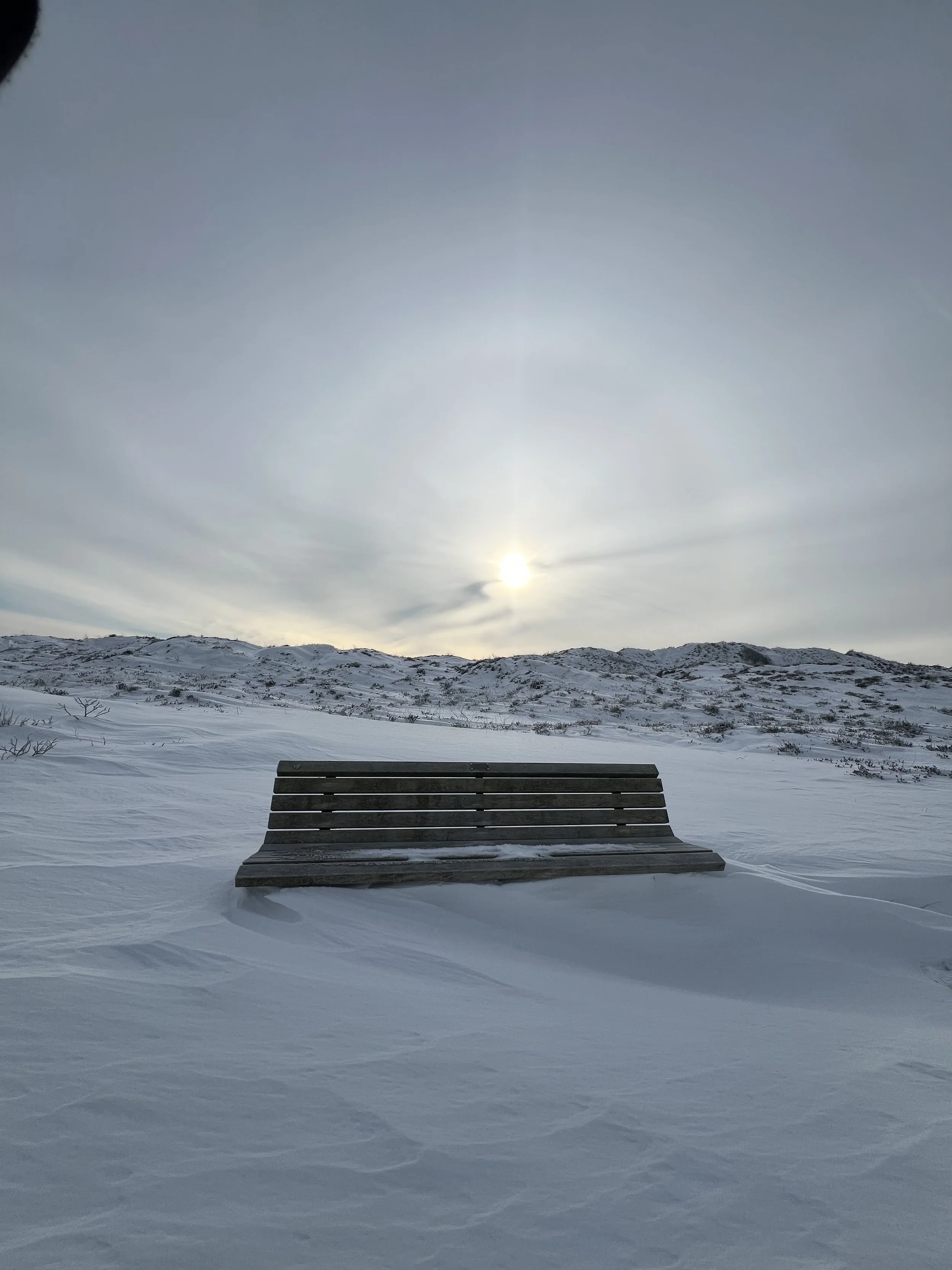 Eine einsame Parkbank im Schnee vor einer schneebedeckten Hügelkette bei bewölktem Himmel, unter einer tiefstehenden Sonne.
