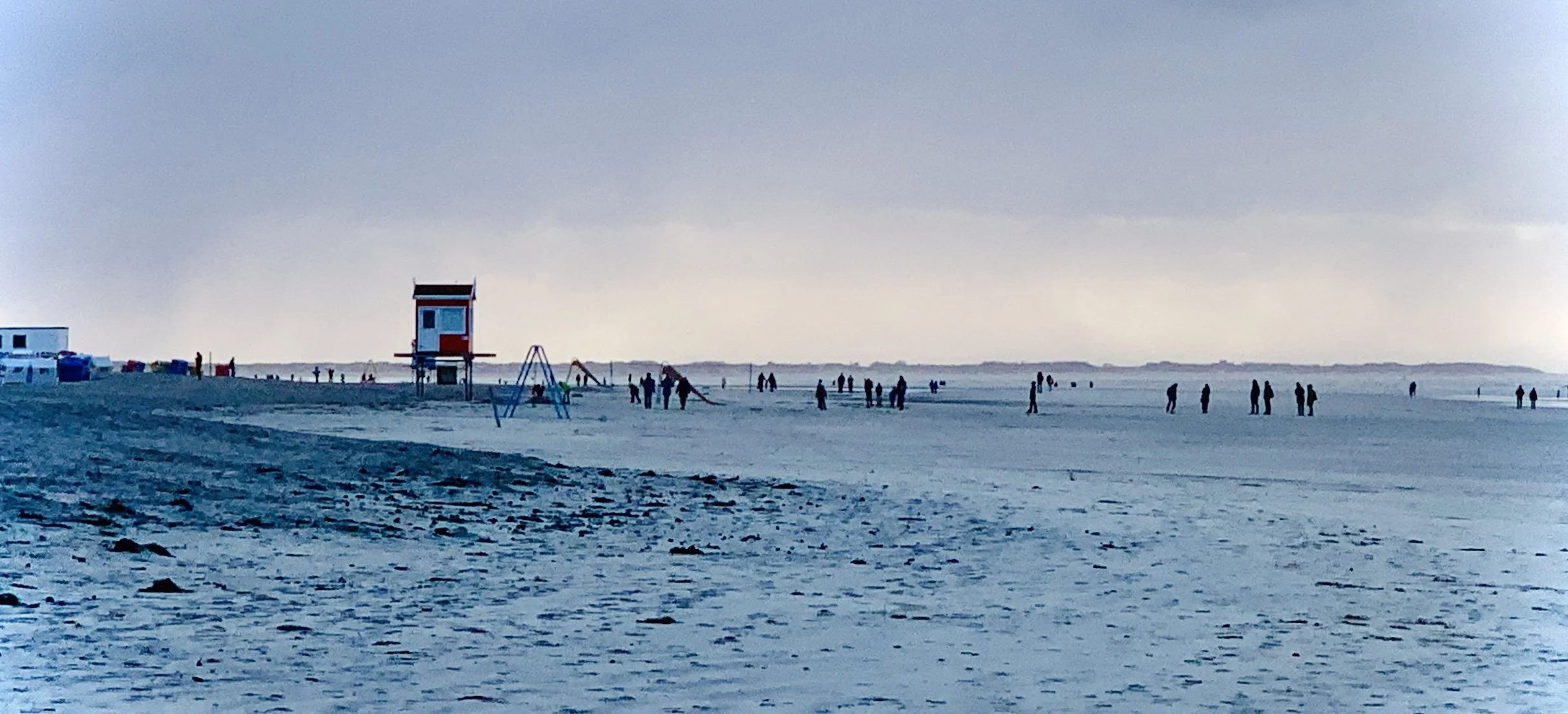 Ein Strand bei Sonnenuntergang mit mehreren Personen, einer Rettungststation und Spielgeräten im Hintergrund.