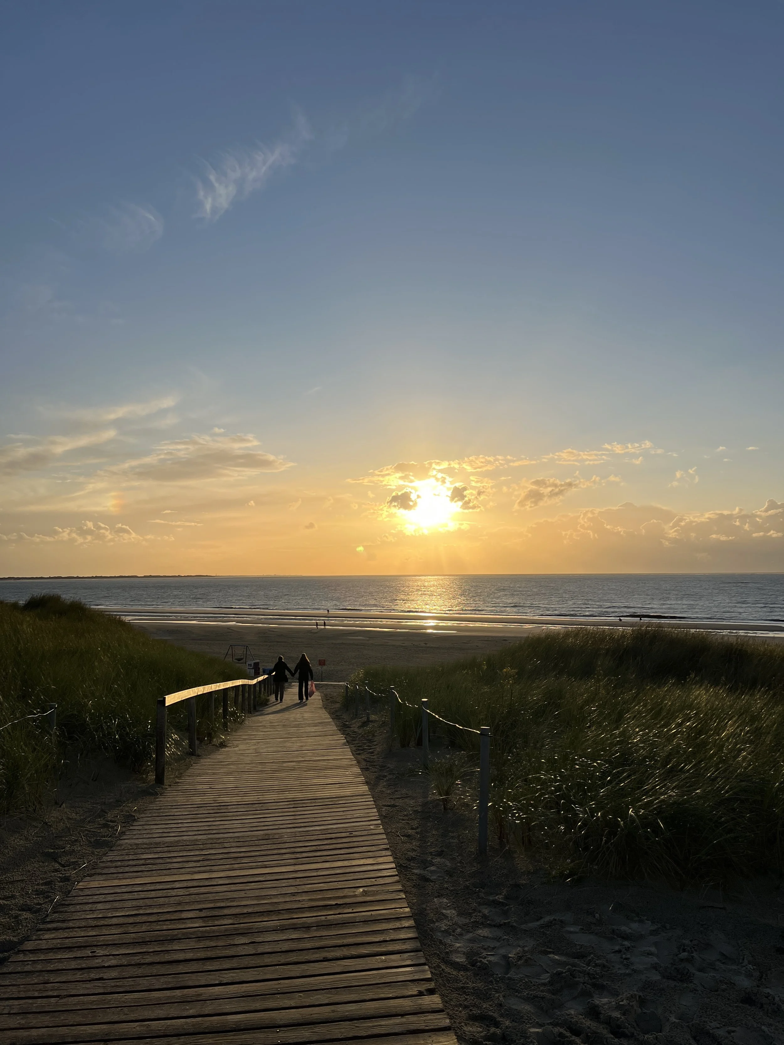 Ein Holzsteg führt durch Sanddünen zum Strand bei Sonnenuntergang, mit zwei Personen, die Hand in Hand gehen, und dem Meer im Hintergrund.