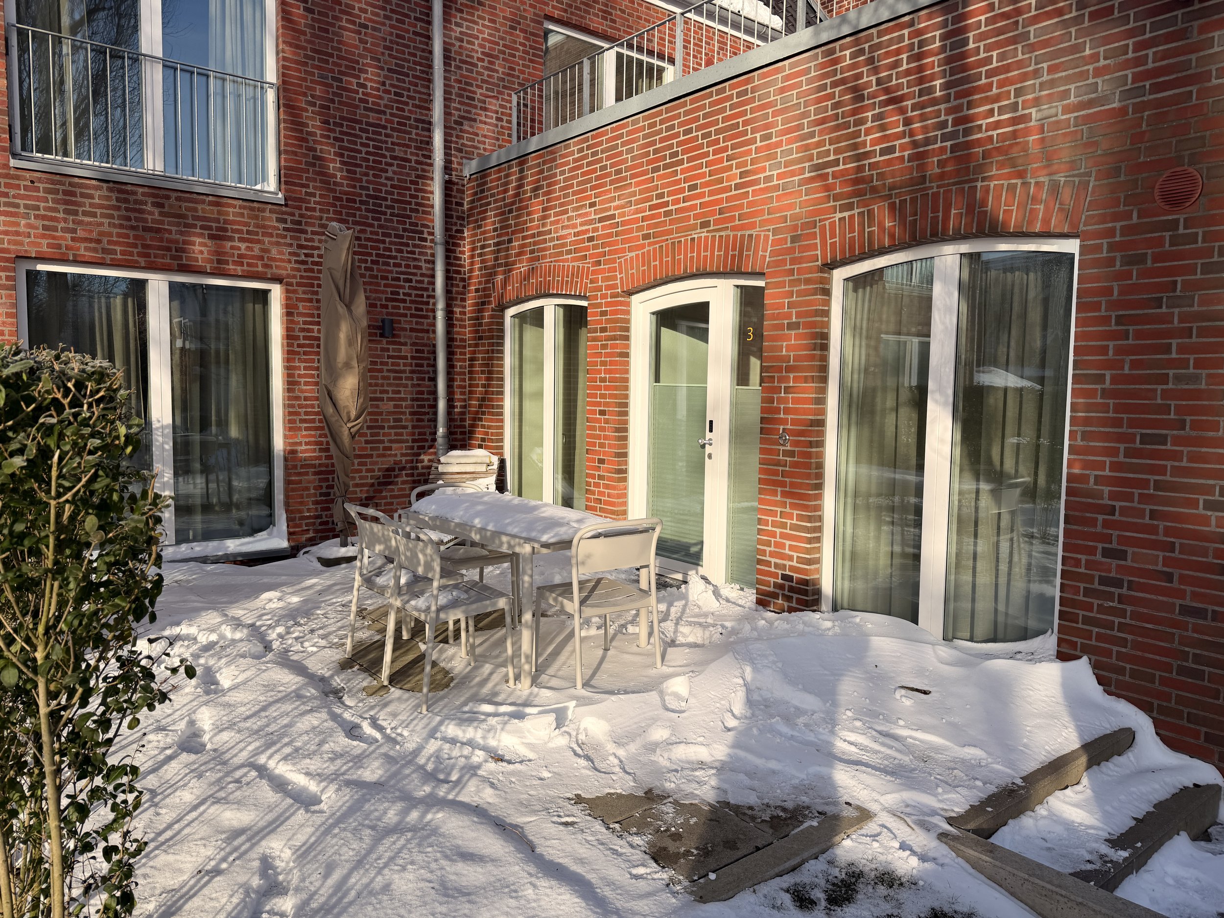 Ein schneebedeckter Hof mit einem runden Tisch, vier Stühlen, einem Sonnenschirm, einer Tür mit der Nummer 3, und großen Fenstern an einer roten Backsteinwand.