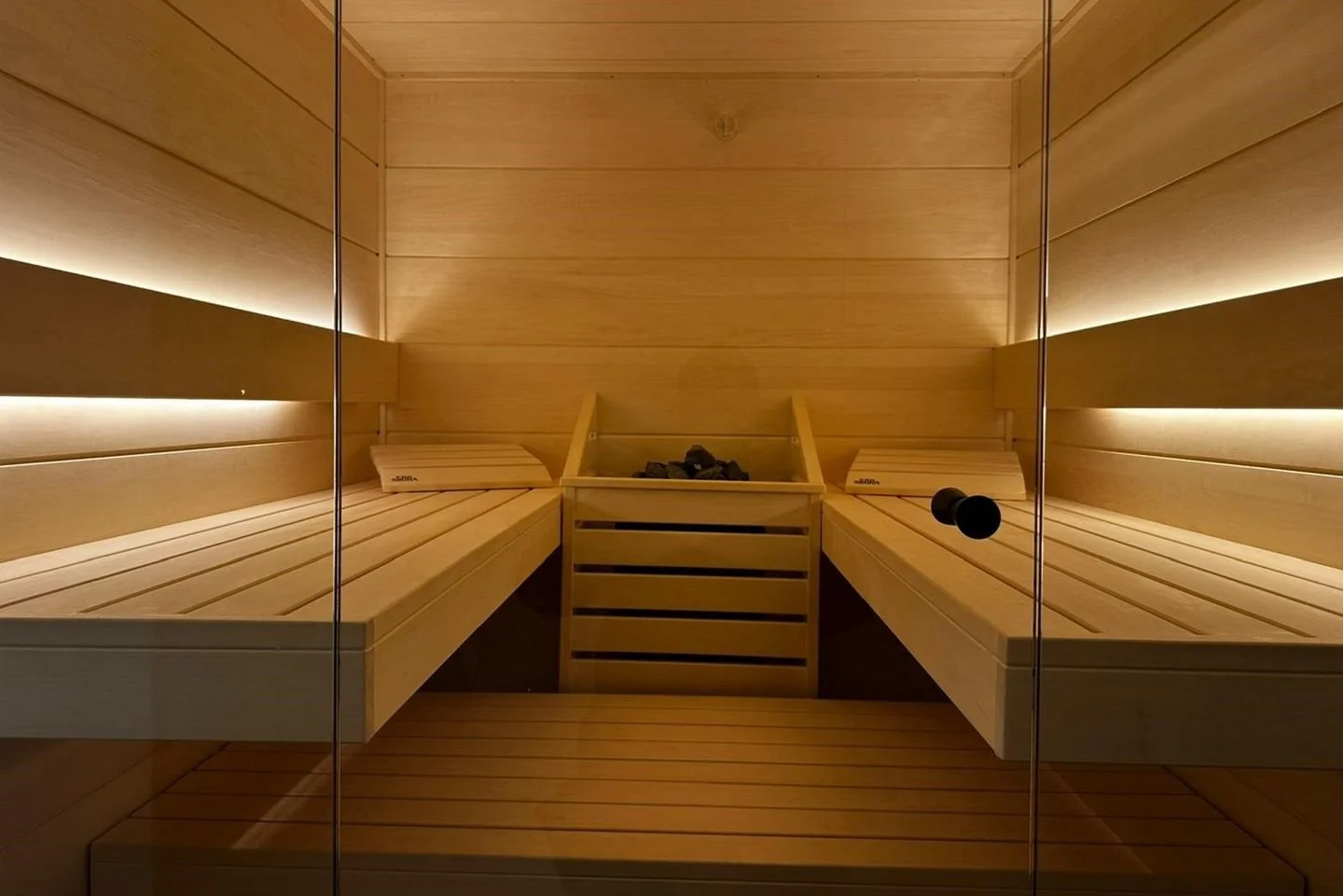 Innenansicht einer modernen Sauna mit Holzpolstern, Steinen in der Saunakiste und einer Glastür.