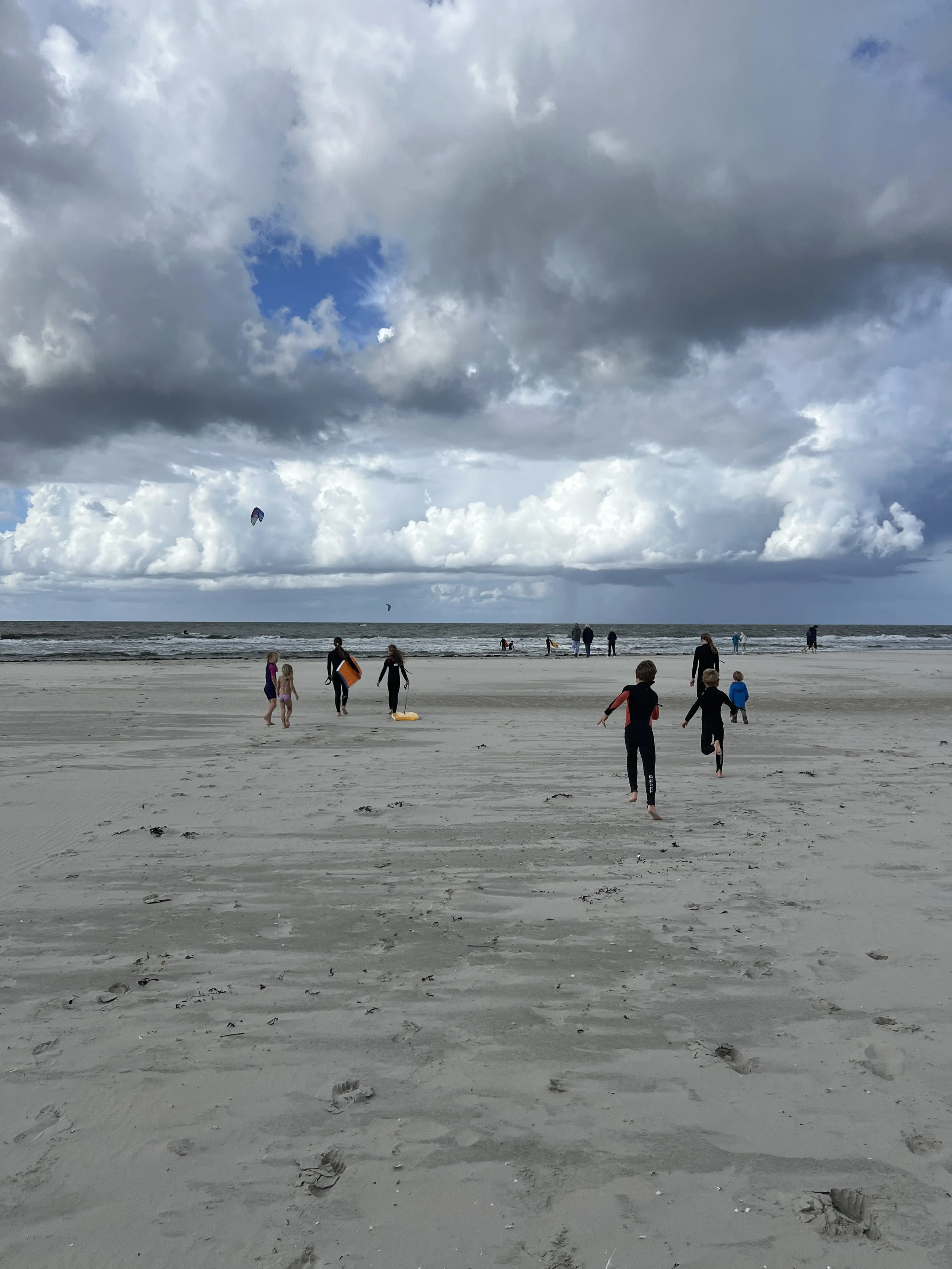 Kinder und Erwachsene am Strand bei bewölktem Himmel, einige Kitesurfer im Wasser.