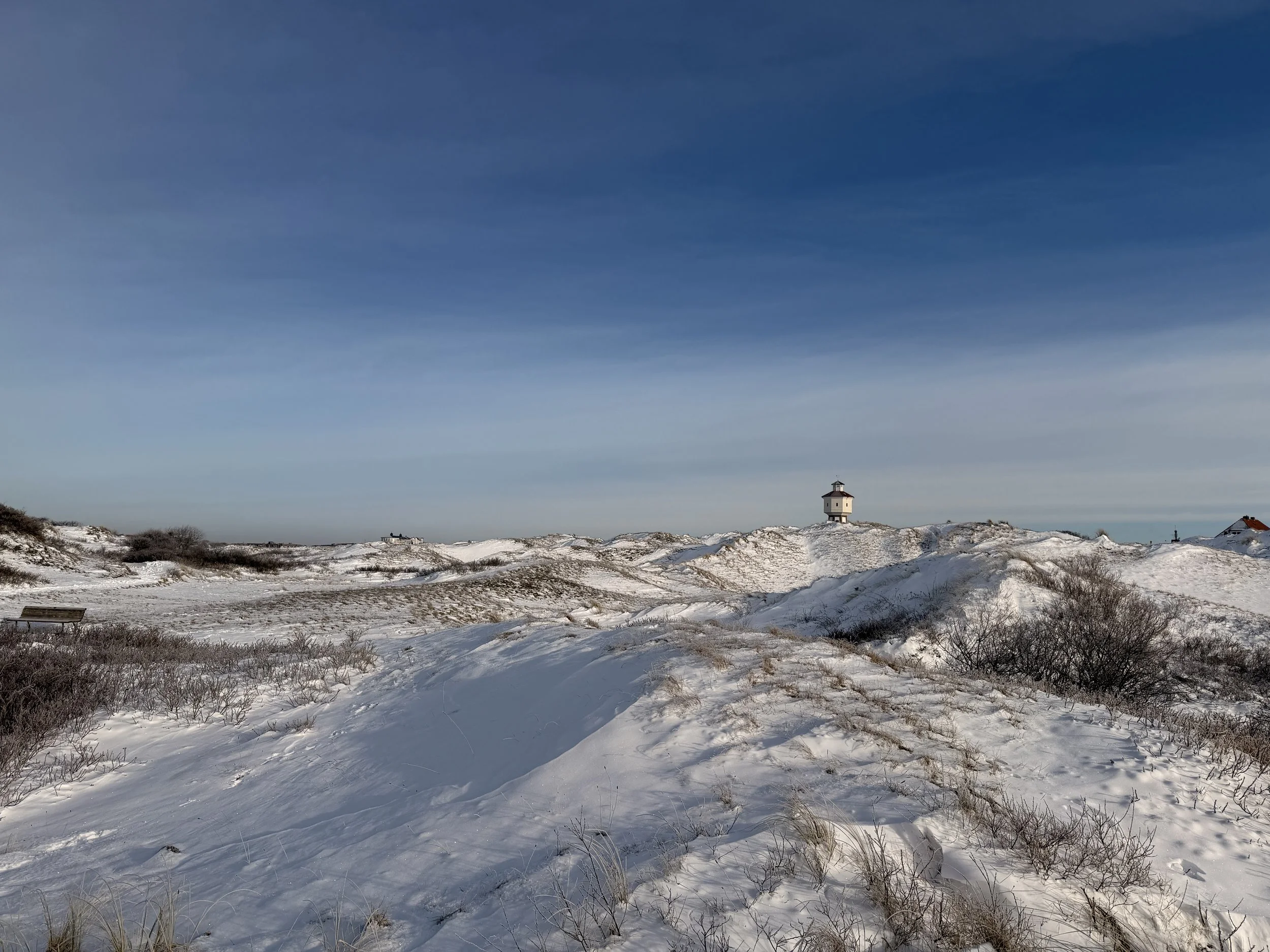 Schneebedeinte Dünenlandschaft mit einem Leuchtturm auf einem Hügel und einem blauen Himmel.