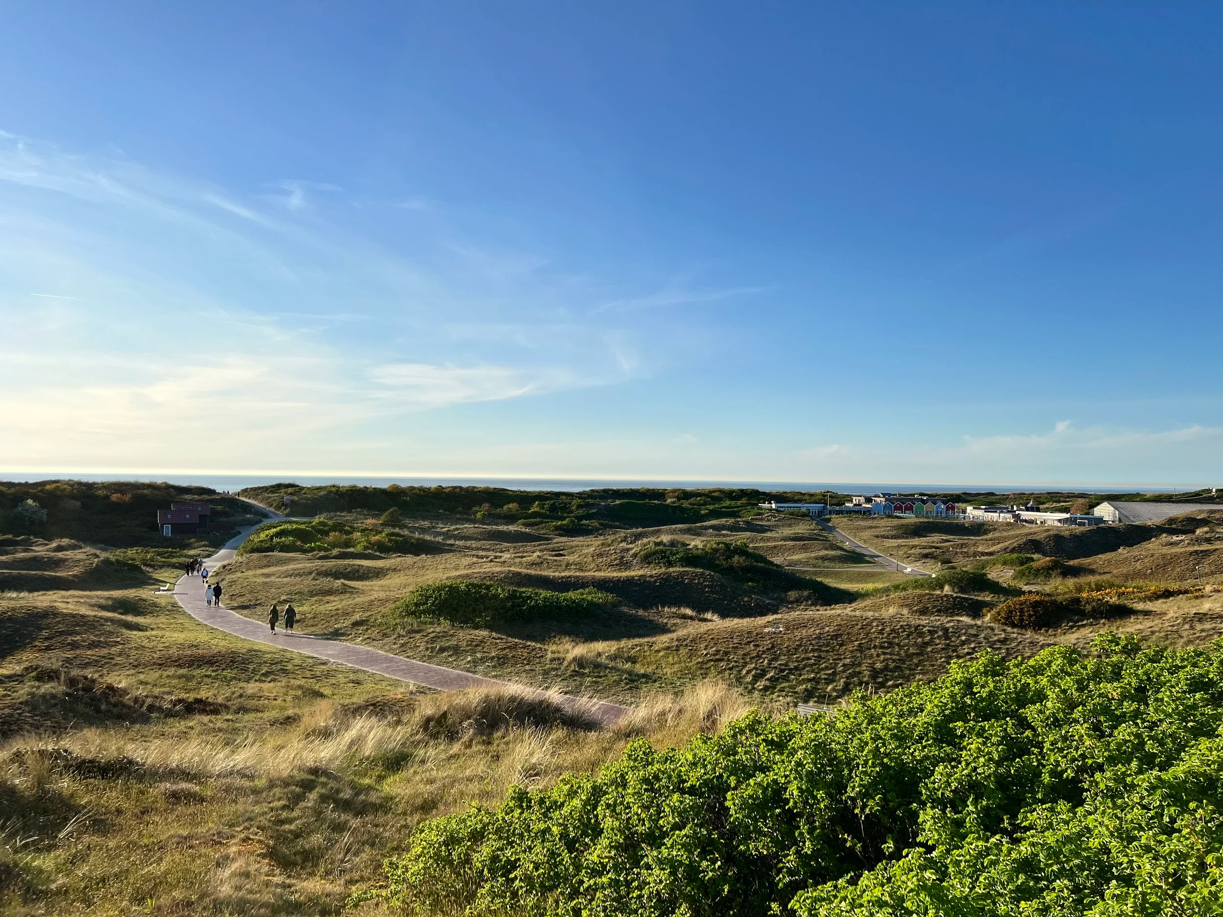 Eine offene Landschaft mit sanften Hügeln, einem Gehweg, ein paar Spaziergängern und Gebäuden im Hintergrund, bei sonnigem Himmel.