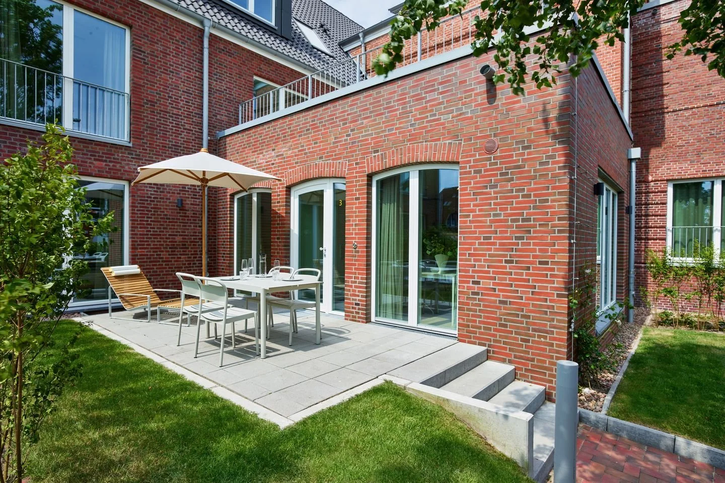 Ein rotes Backsteinhaus mit einem Garten und einer Terrasse, auf der ein Esstisch mit Stühlen und einem Sonnenschirm steht.