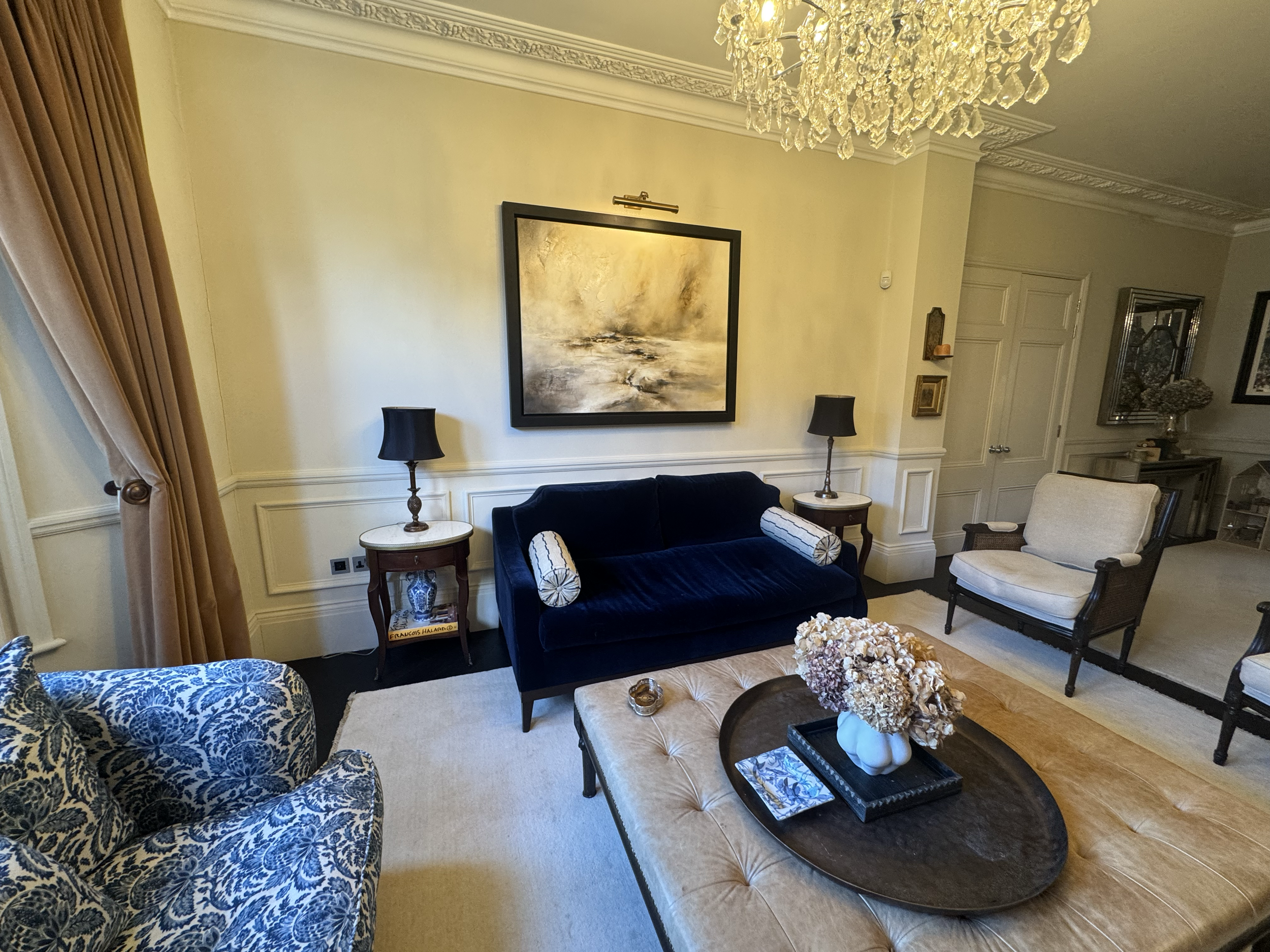 Living room with a blue velvet loveseat, beige armchairs, side tables with black lamps, a large abstract painting on the wall, a chandelier, and a table with a floral arrangement.