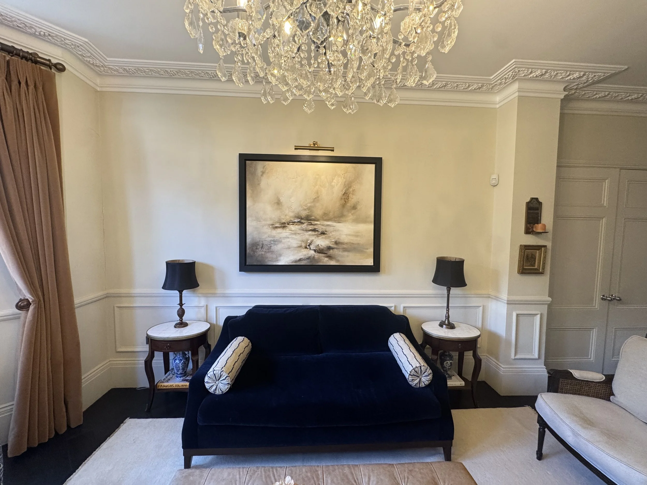 Living room with a black velvet sofa, two side tables with black lamps, a beige armchair, a large abstract painting above the sofa, a chandelier hanging from the ceiling, and beige walls with decorative molding.