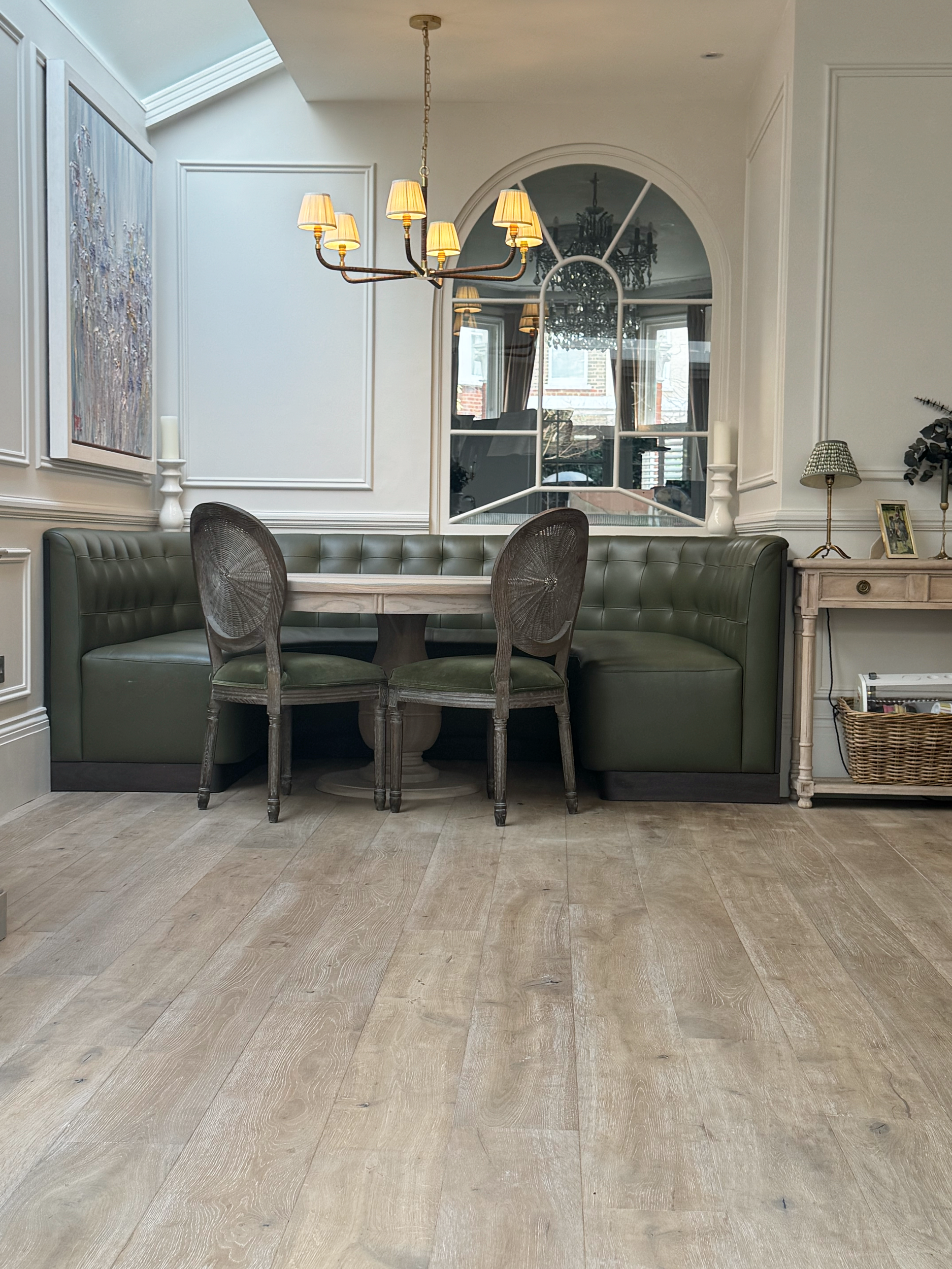 Dining area with a round table, green upholstered banquette, two wooden chairs, chandelier, large arched window, and wall decorations in a cozy, well-lit room.