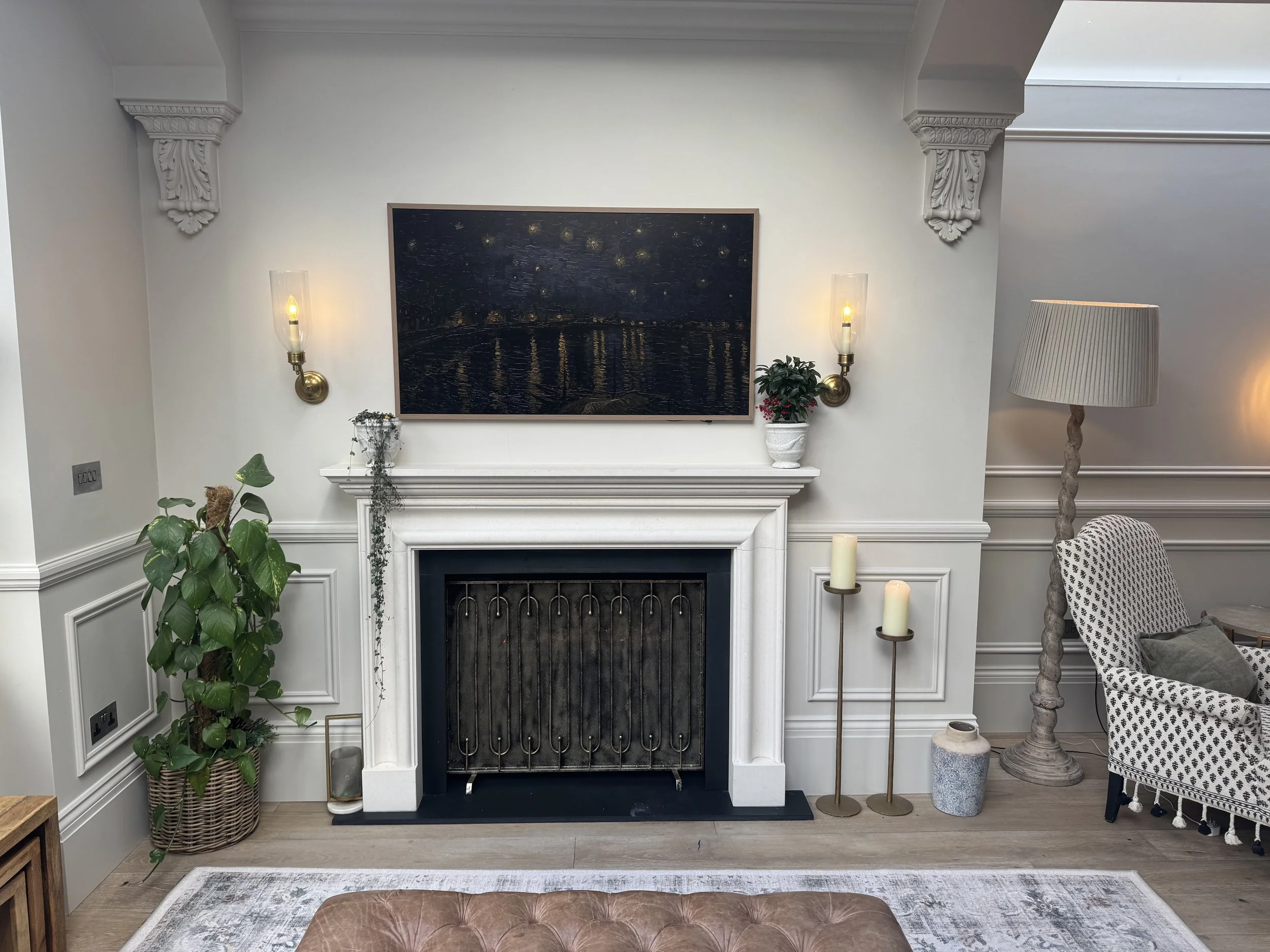 Living room with a fireplace, framed artwork above, wall sconces, potted plant, candles, floor lamp, patterned armchair, and ottoman.