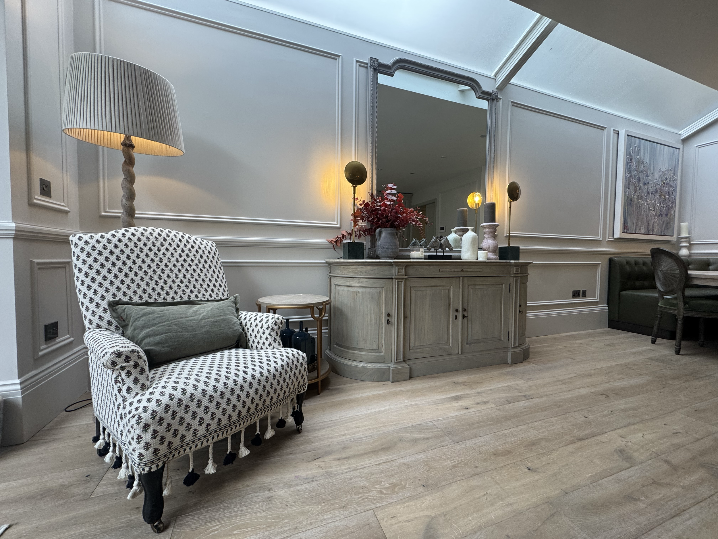 Interior of a living room with a white armchair, a tall floor lamp, a side table, a console table with decorative vases and artwork, and a green cushioned seating area.