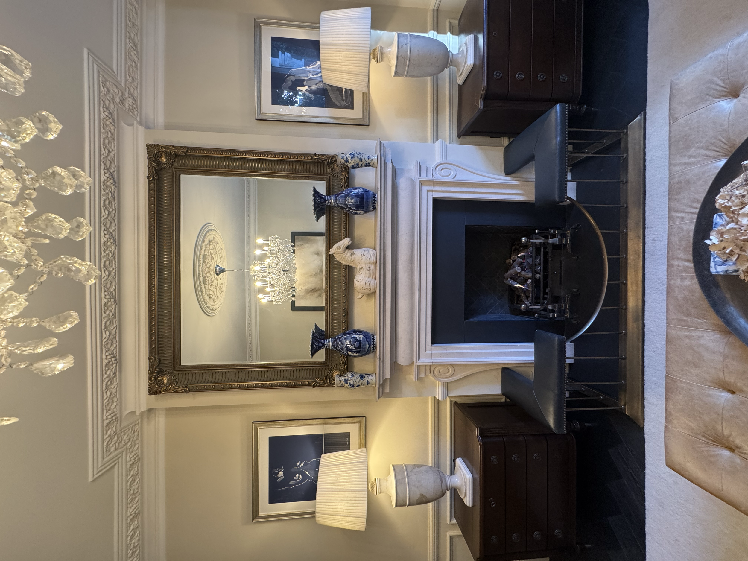 Elegant living room with a fireplace, large framed mirror, two table lamps, decorative vases, chandelier, and artwork on the walls.