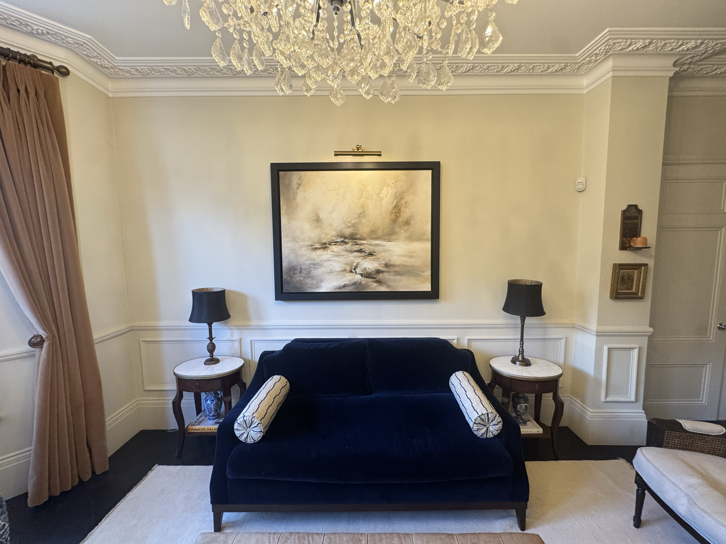 Living room with a black velvet sofa, two white side tables with black lamps, a chandelier, beige walls, and a framed abstract landscape painting.