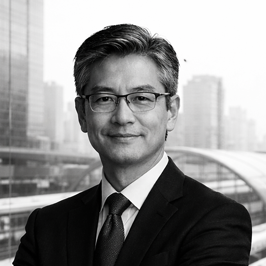 Black and white professional portrait of an Asian man in a suit and glasses, standing with arms crossed in front of a cityscape with modern buildings.