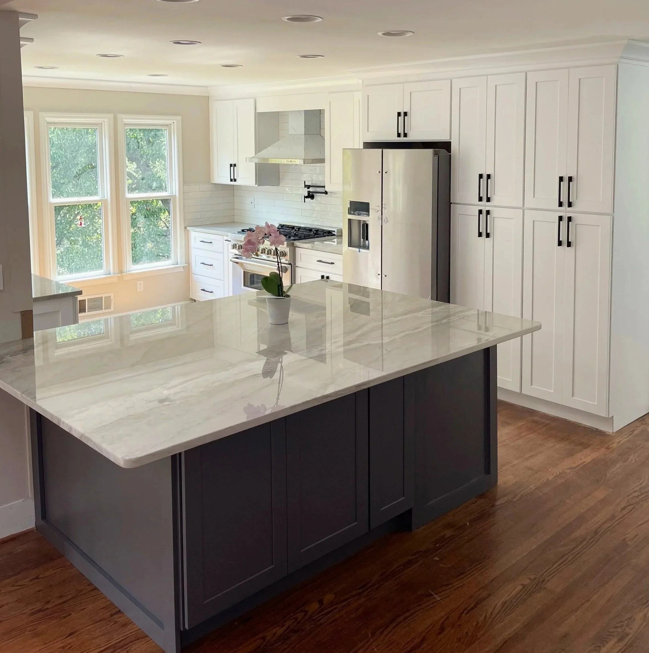 Modern kitchen with white cabinets, stainless steel refrigerator, marble island, and wooden floor, with a pink orchid on the island counter.
