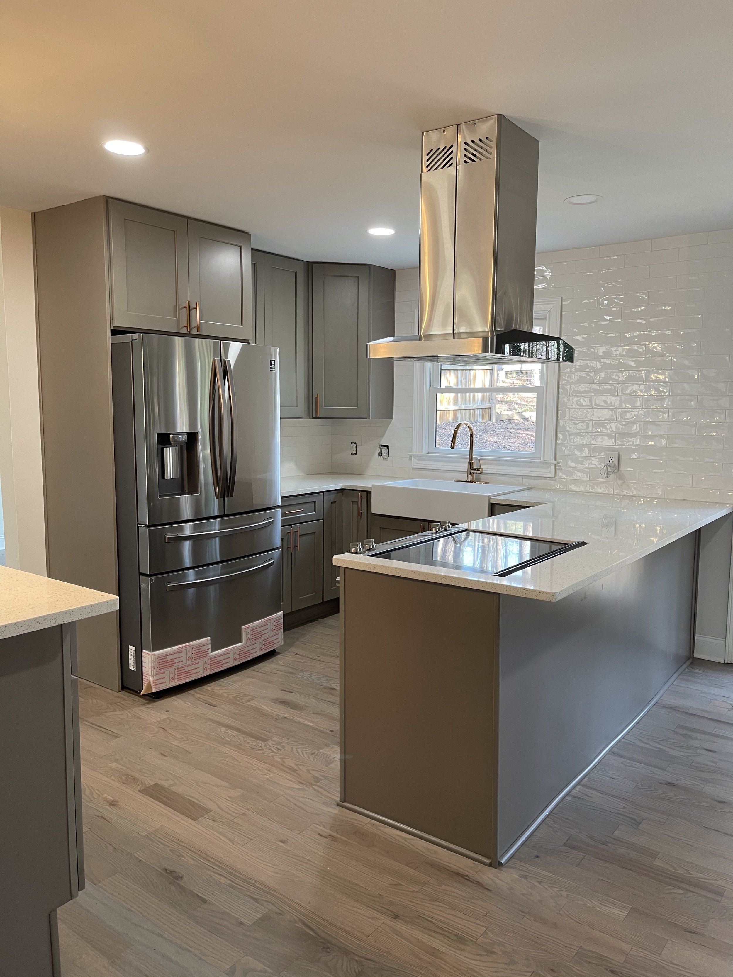 Modern kitchen with gray cabinets, stainless steel refrigerator, island with white countertop, and a range hood over a cooktop.