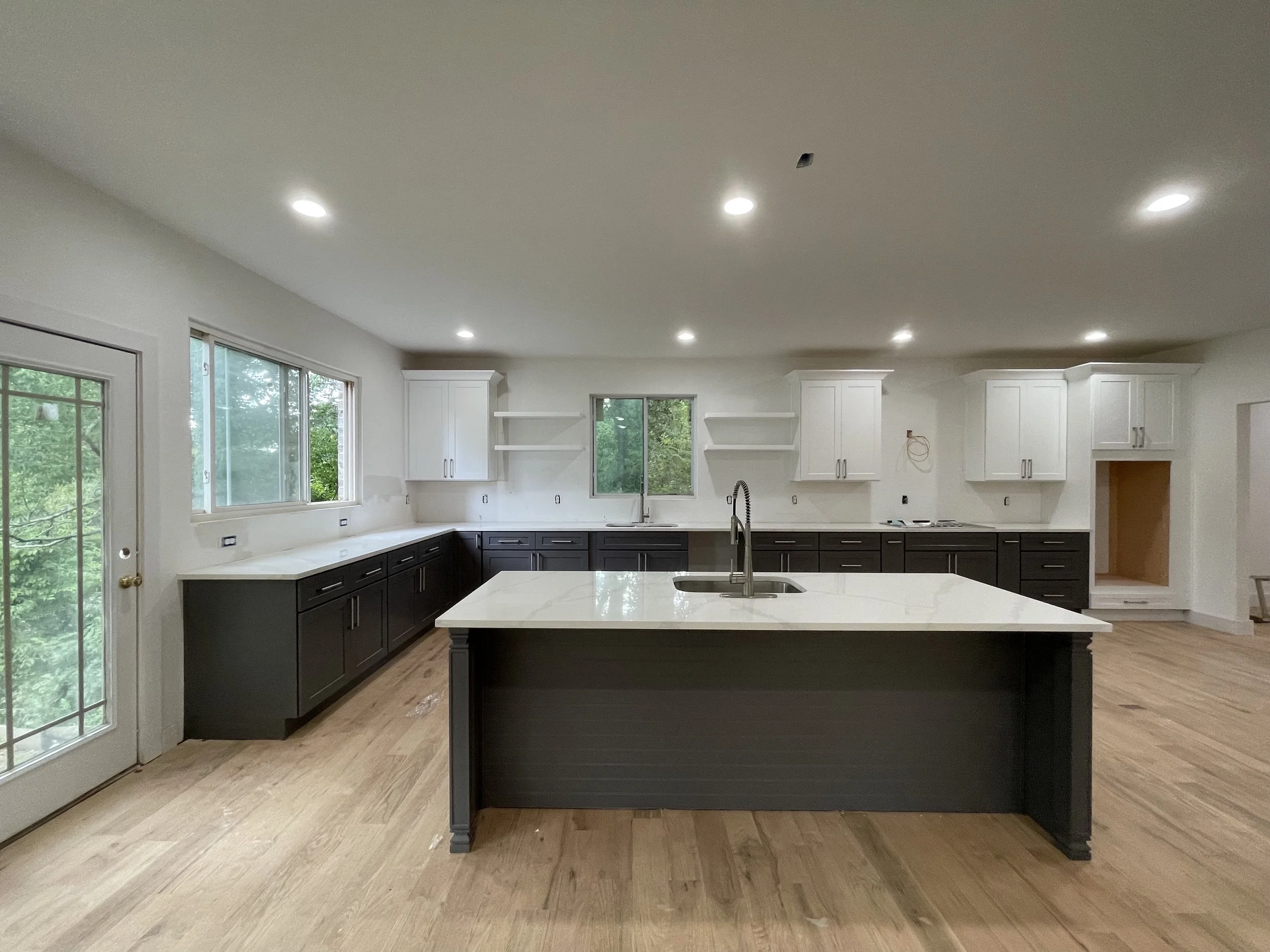 Modern kitchen with white upper cabinets, black lower cabinets, island with white countertop, wooden flooring, windows view of greenery, and recessed ceiling lights.