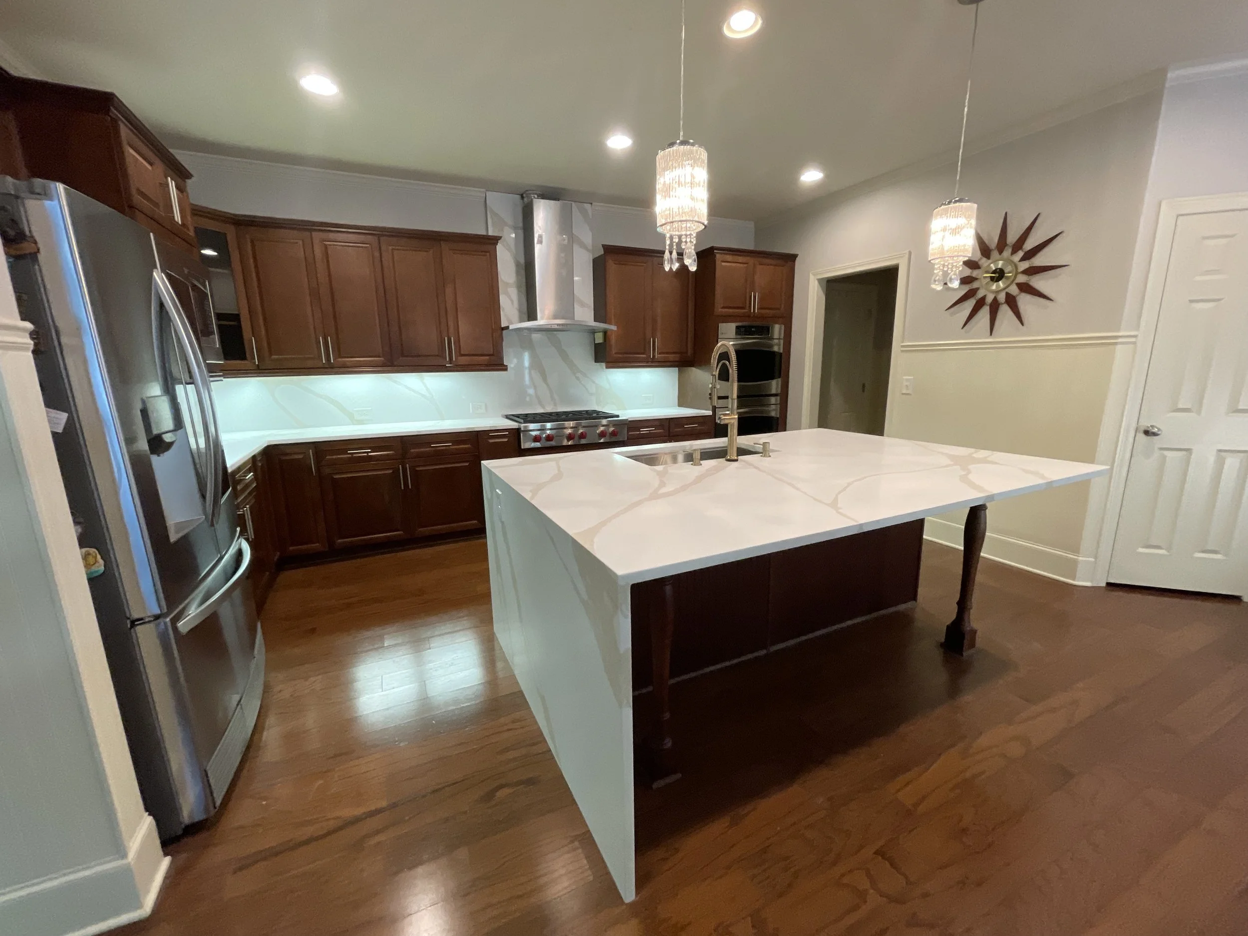 Modern kitchen with wooden cabinets, stainless steel appliances, a white marble island, and hardwood floors, illuminated by recessed and pendant lighting.