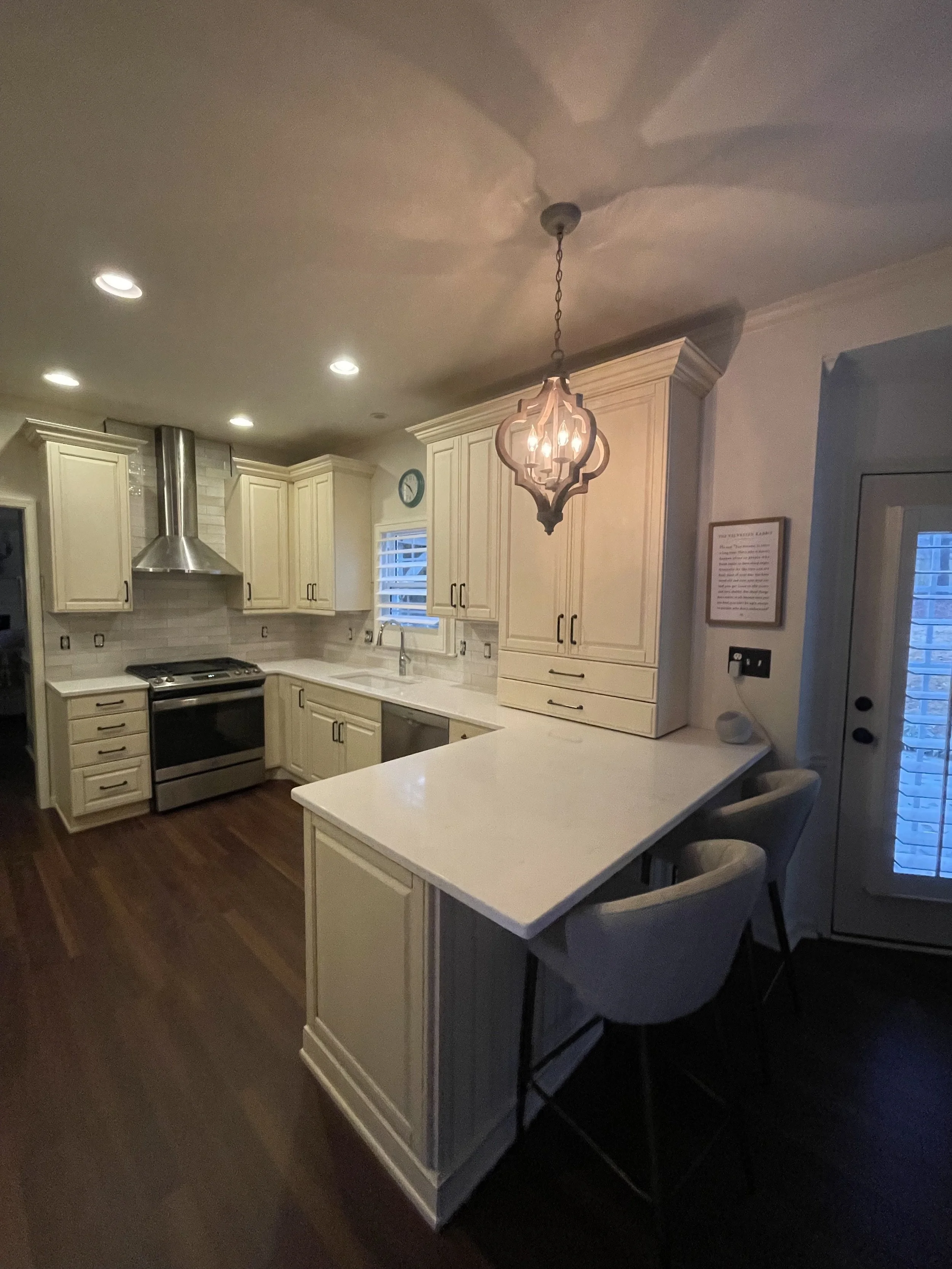 Modern kitchen with white cabinets, stainless steel oven and range, white countertops, hardwood floors, decorative pendant light, window with blinds, and kitchen bar with two chairs.