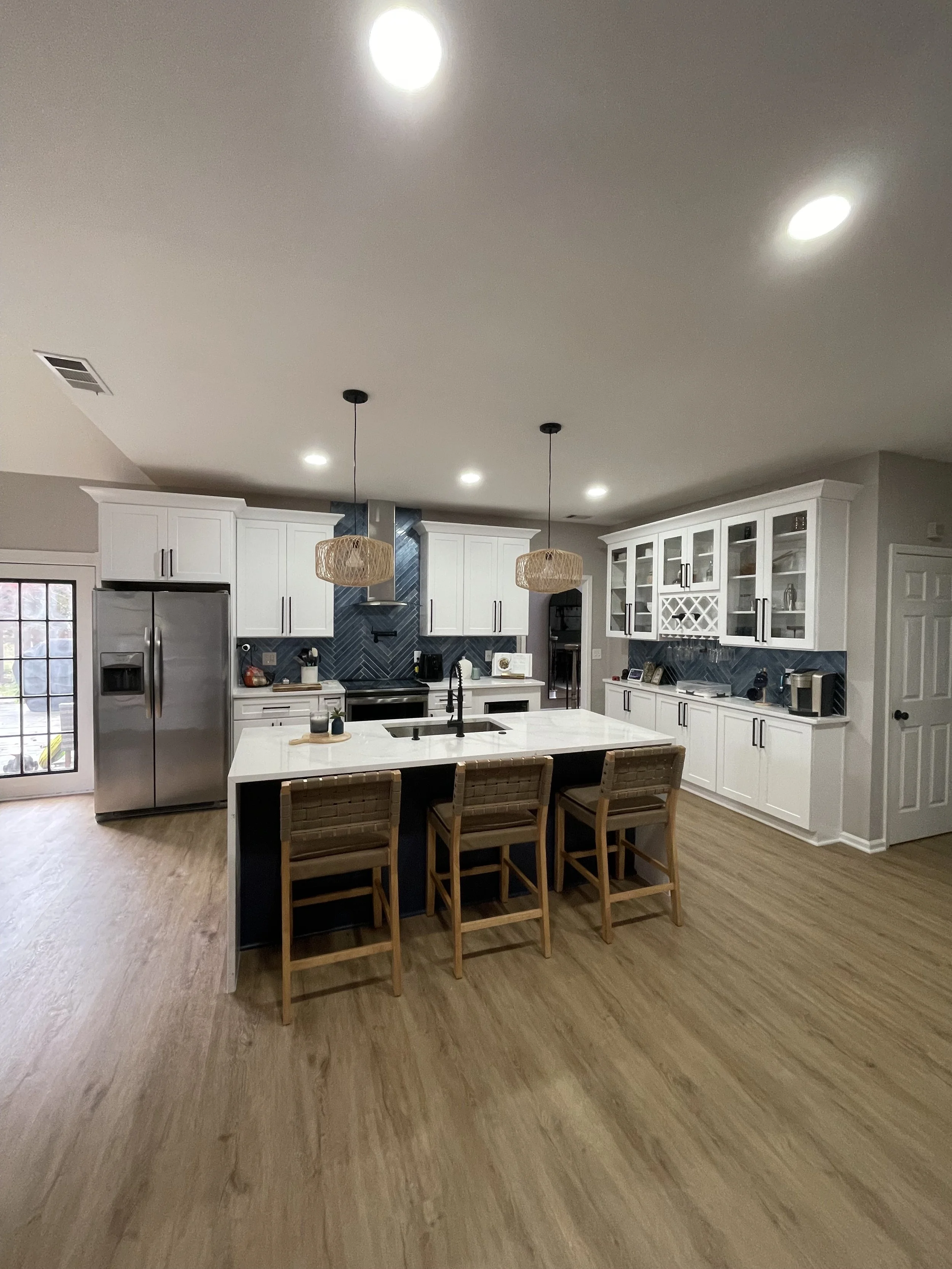 Modern kitchen with white cabinets, a large island with a white countertop and four wooden chairs, dark blue backsplash, stainless steel appliances, wooden flooring, and pendant lights hanging over the island.