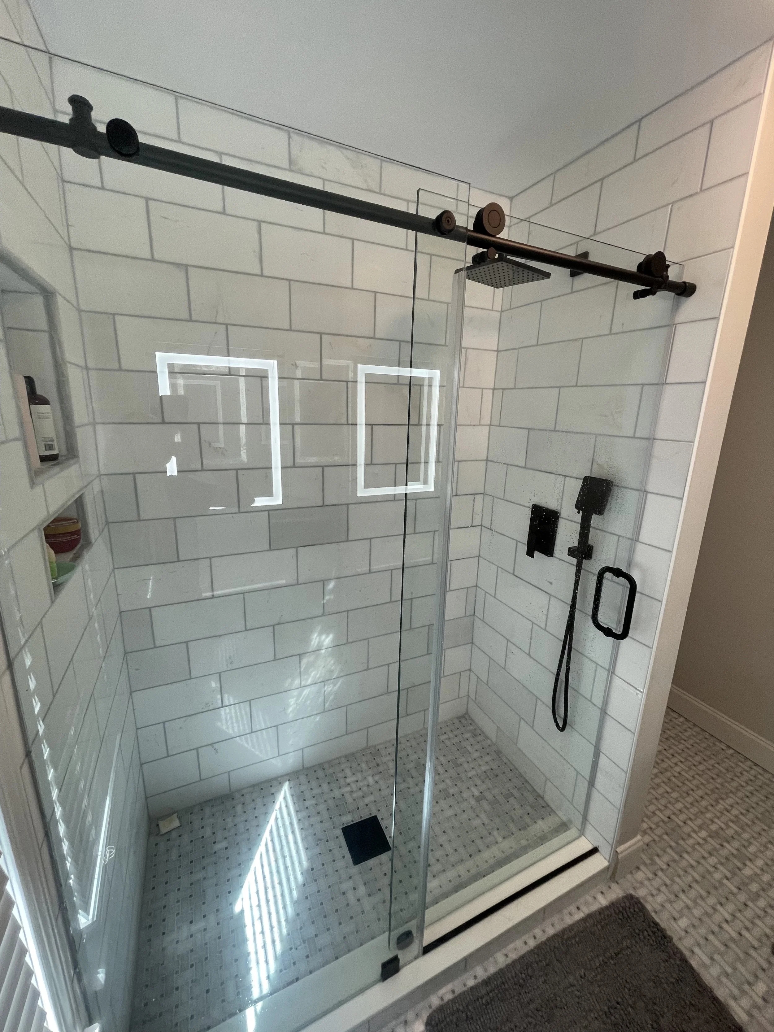 A modern shower with clear glass doors, white subway tile walls, and a black rain showerhead mounted on a black sliding bar. A handheld showerhead is attached on the right wall, and there's a built-in niche with toiletries on the left wall. The floor