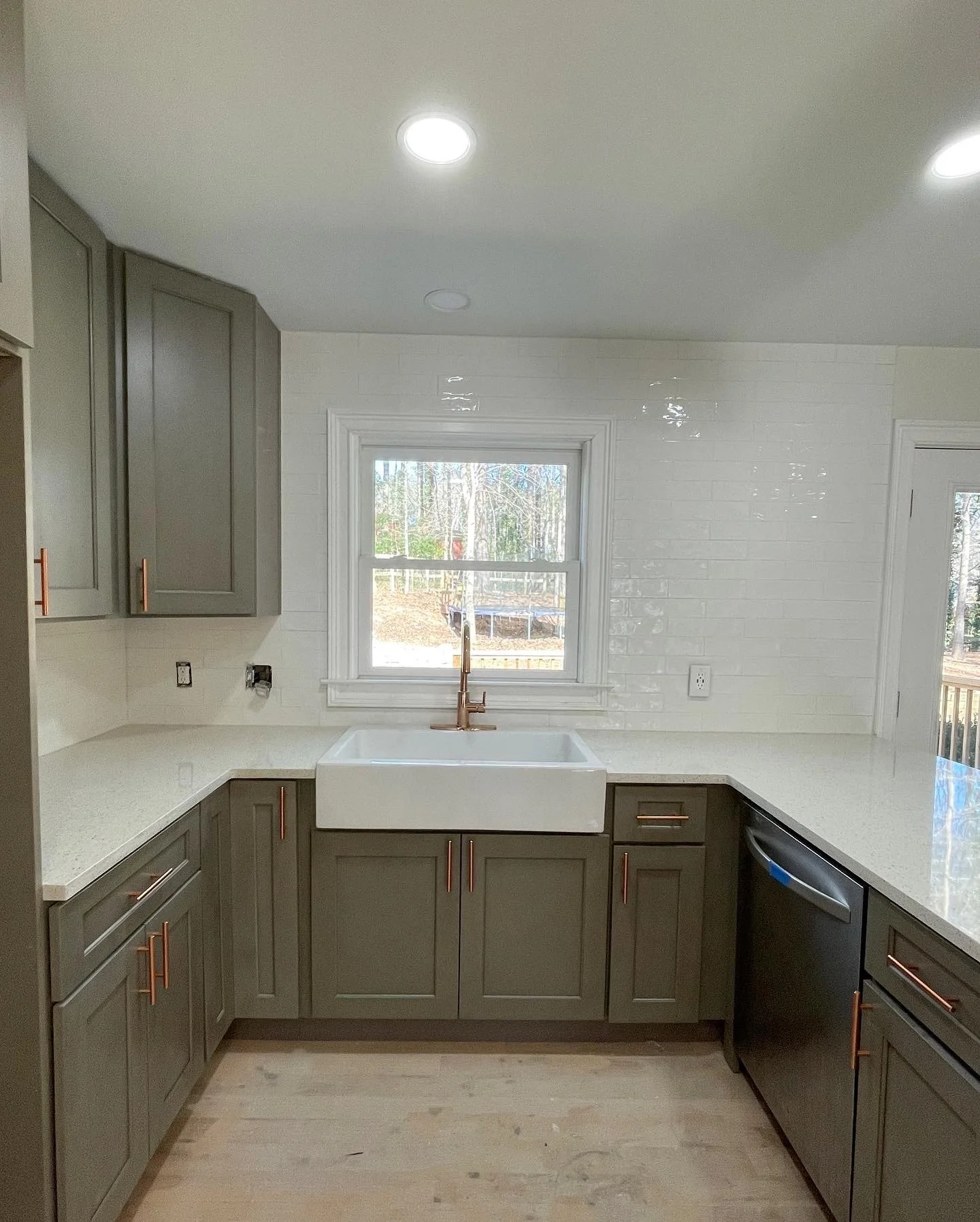 Modern kitchen with gray cabinets, white countertops, a farmhouse-style sink, and a window over the sink. Recessed lighting and hardwood floors are visible.