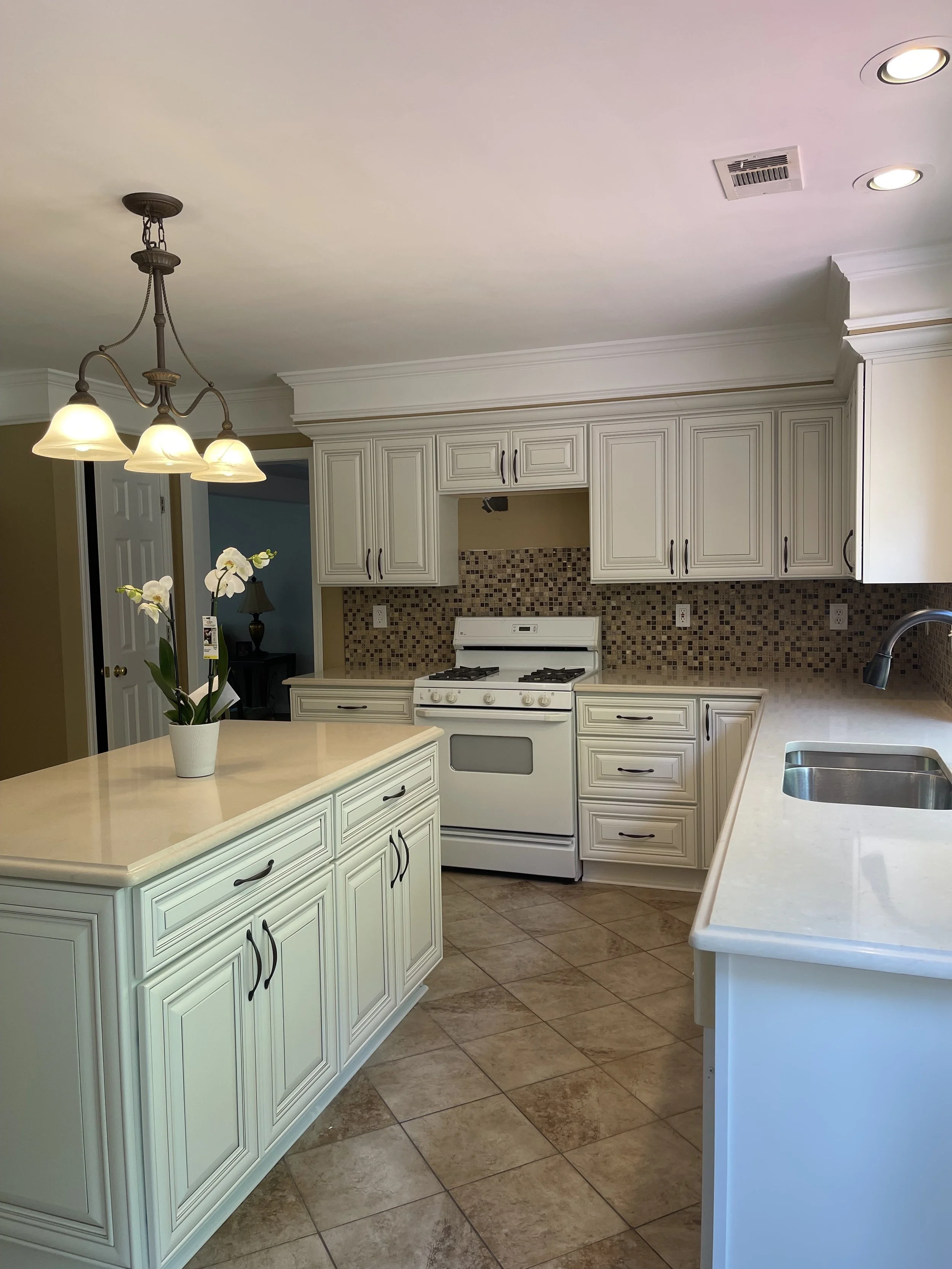 A clean, white kitchen with an island, a stove, and a beige tiled backsplash. A potted white orchid is on the island, and there are recessed ceiling lights.
