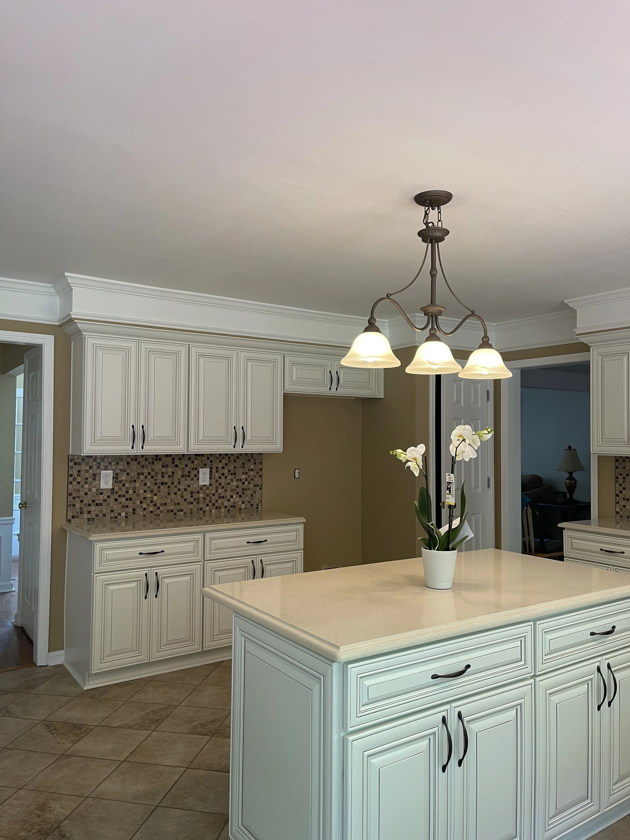 A kitchen with white cabinets, beige walls, a central island with a cream-colored countertop, a small potted orchid on the island, and a chandelier with three lamps hanging from the ceiling.