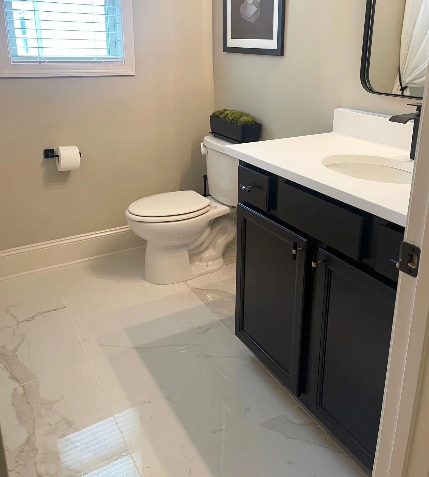 A modern bathroom with beige walls, a white toilet, a black vanity with a white countertop, a round sink, a black framed mirror, a window with blinds, and a small plant on the toilet tank.