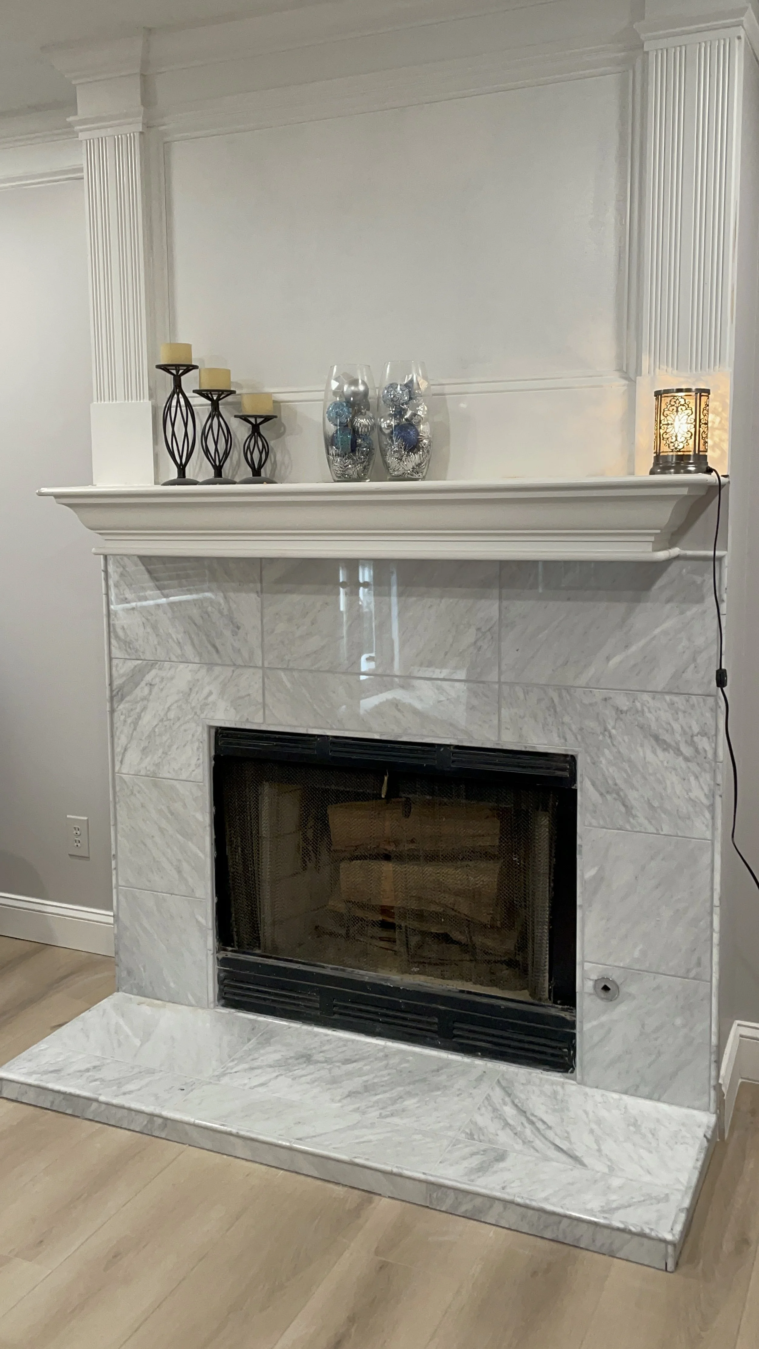 White marble fireplace with decorative items on the mantel, including candle holders with candles, glass vases with blue and silver ornaments, and a small lamp, in a room with light-colored walls and wooden flooring.
