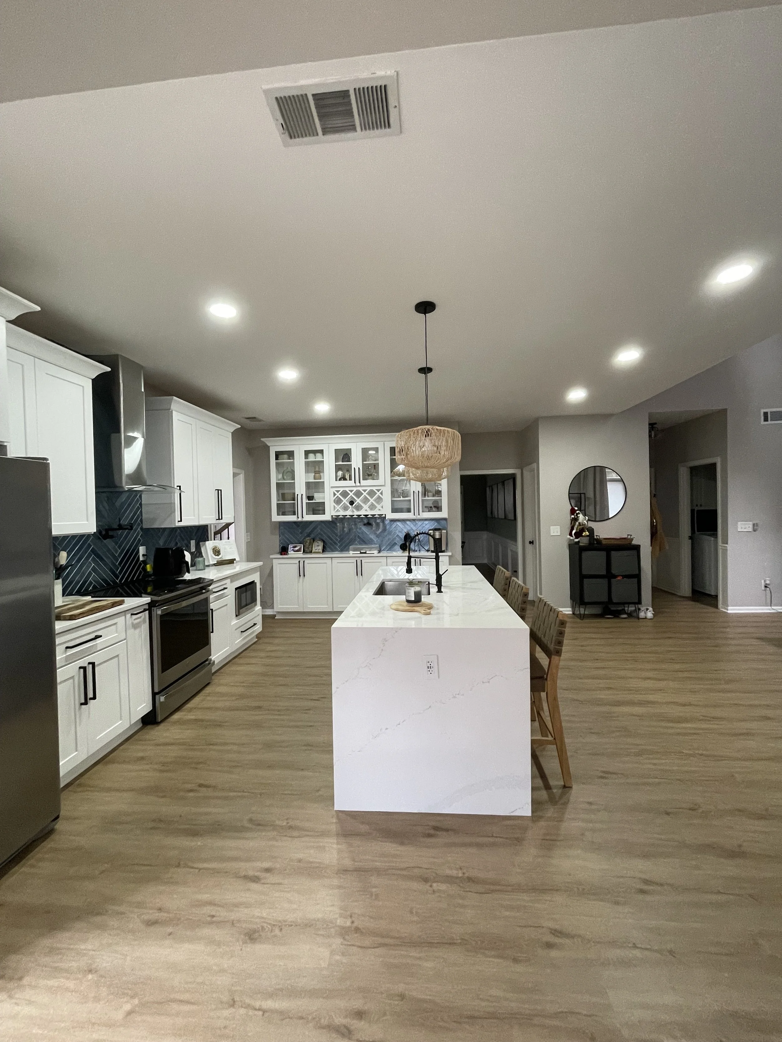Modern kitchen with white cabinets, stainless steel appliances, a large white island with a built-in sink, wooden bar stools, and a decorative light fixture hanging overhead.