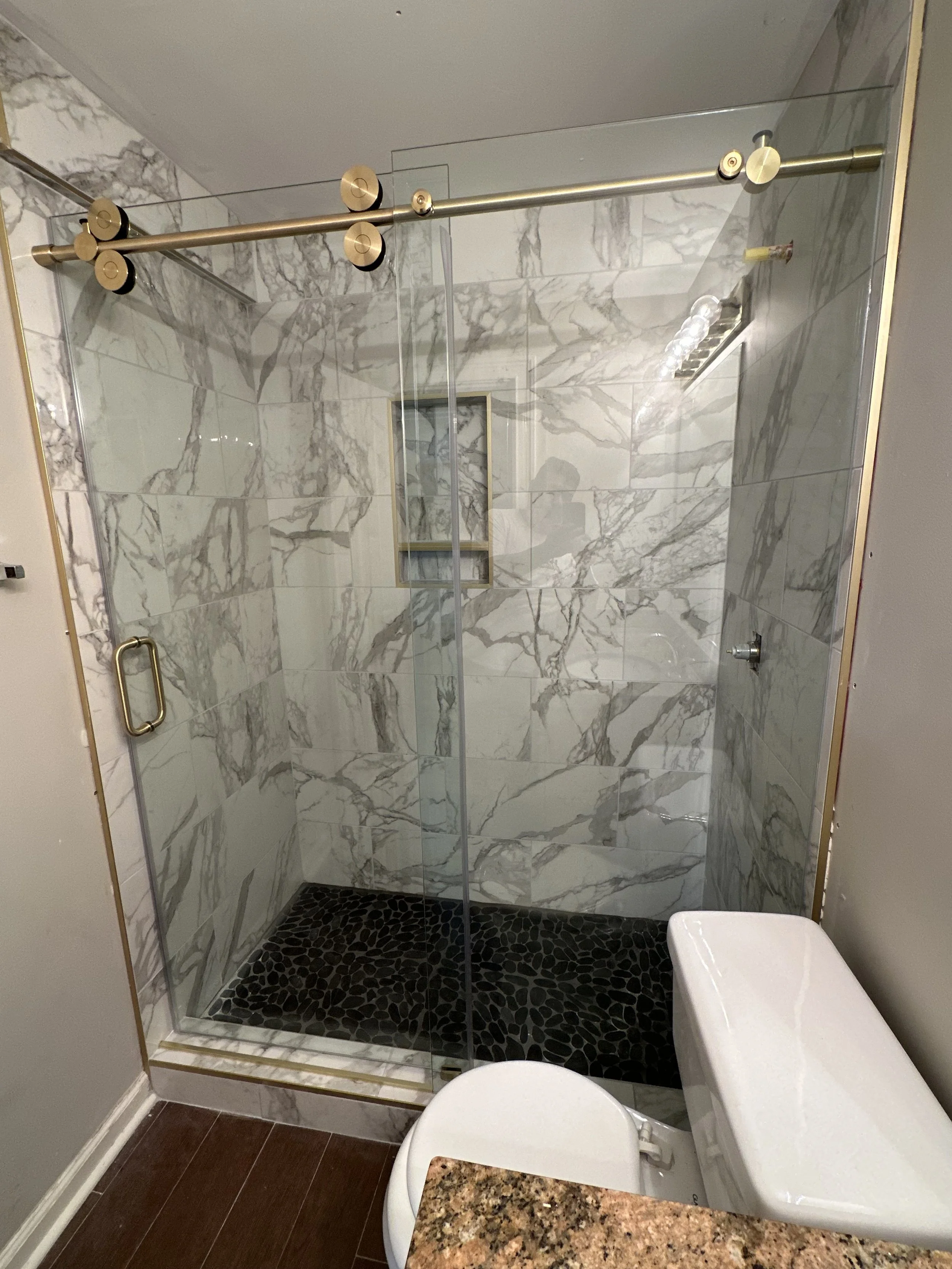Marble-finished walk-in shower with a glass door and a small niche for toiletries. The shower has a black pebble tile floor. A toilet and granite countertop are visible outside the shower.