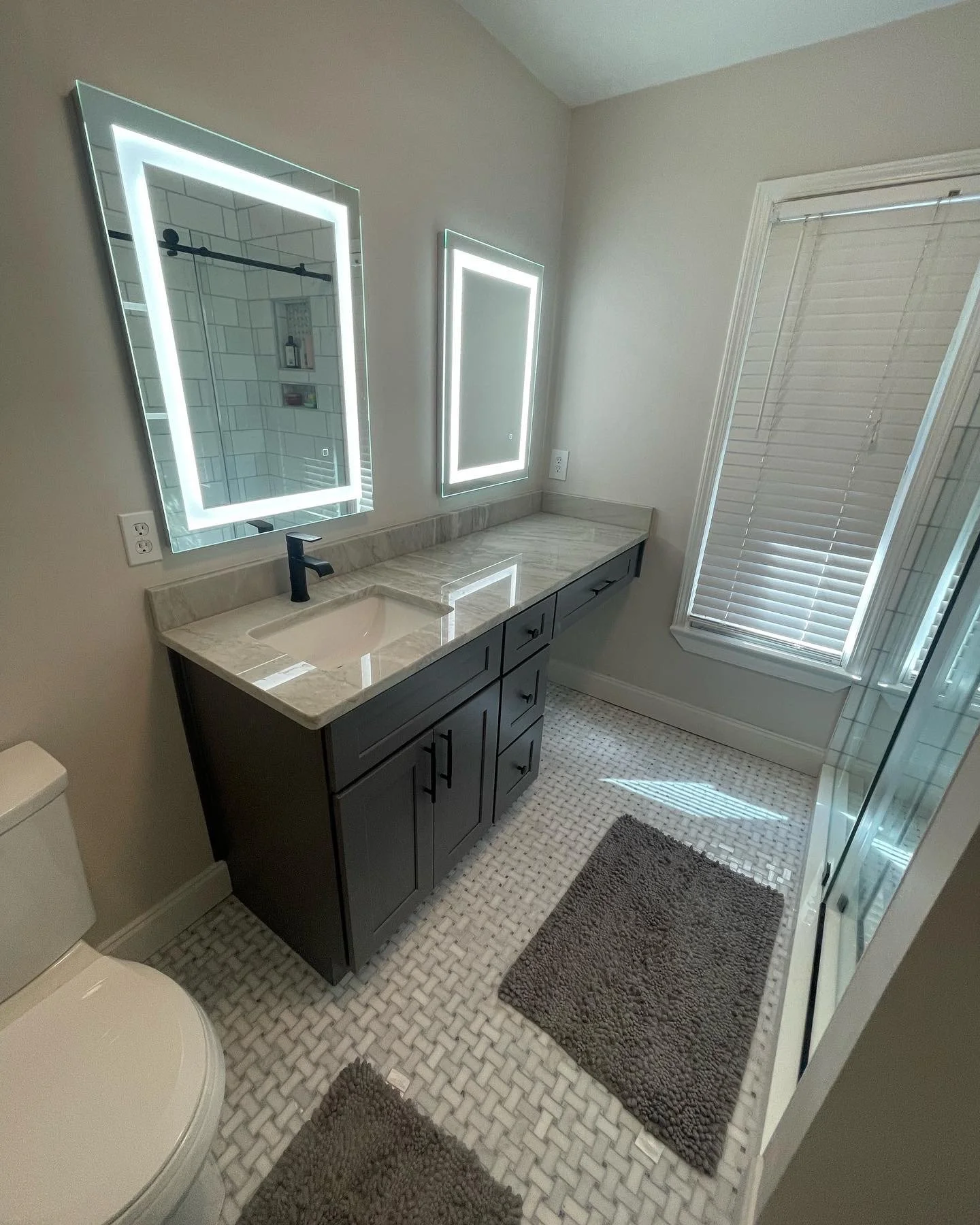 A modern bathroom with a marble countertop vanity, two illuminated rectangular mirrors, a window with blinds, and a walk-in shower with glass doors.