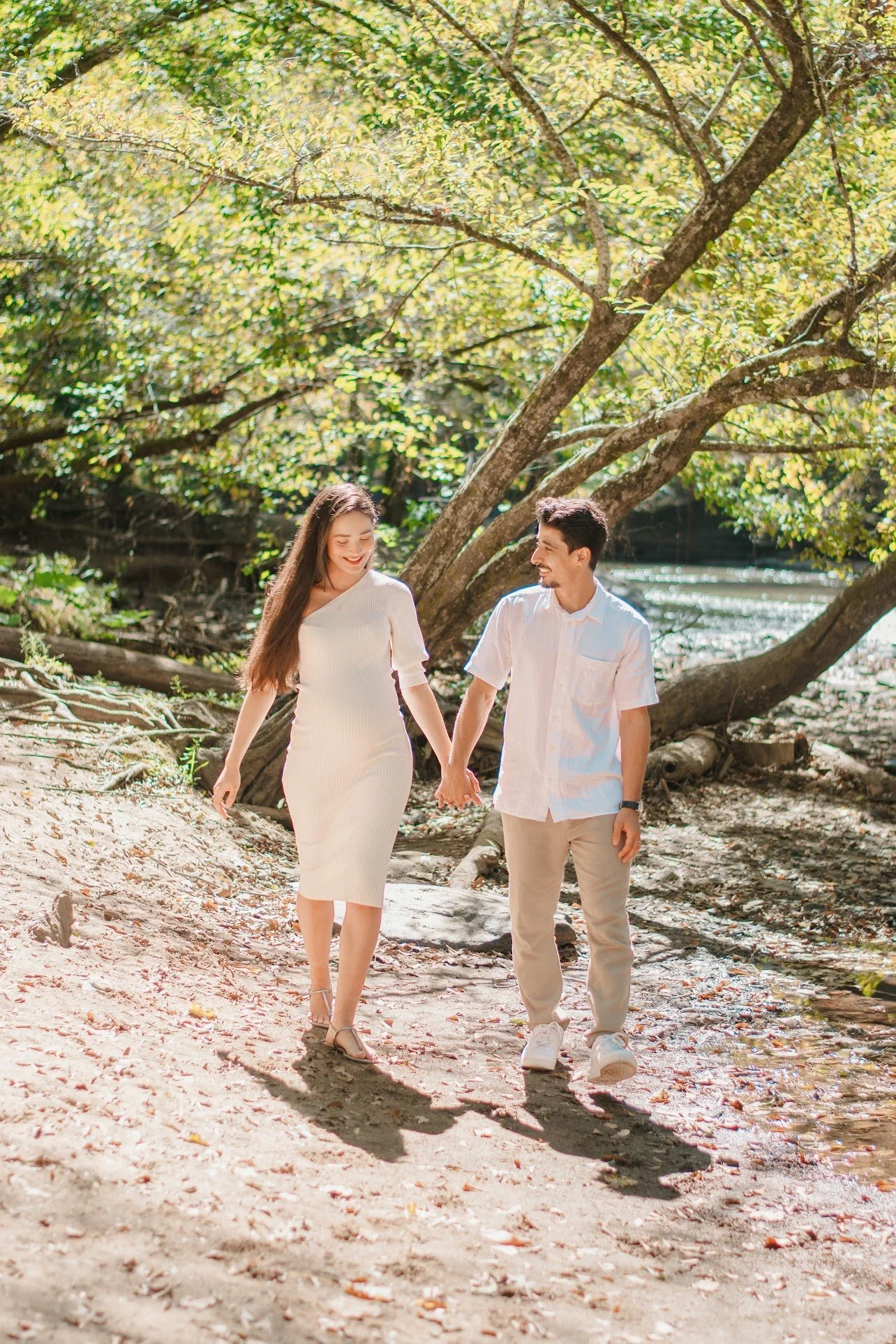A young couple walking hand in hand through a sunlit forested area with a stream nearby, smiling and enjoying each other's company.