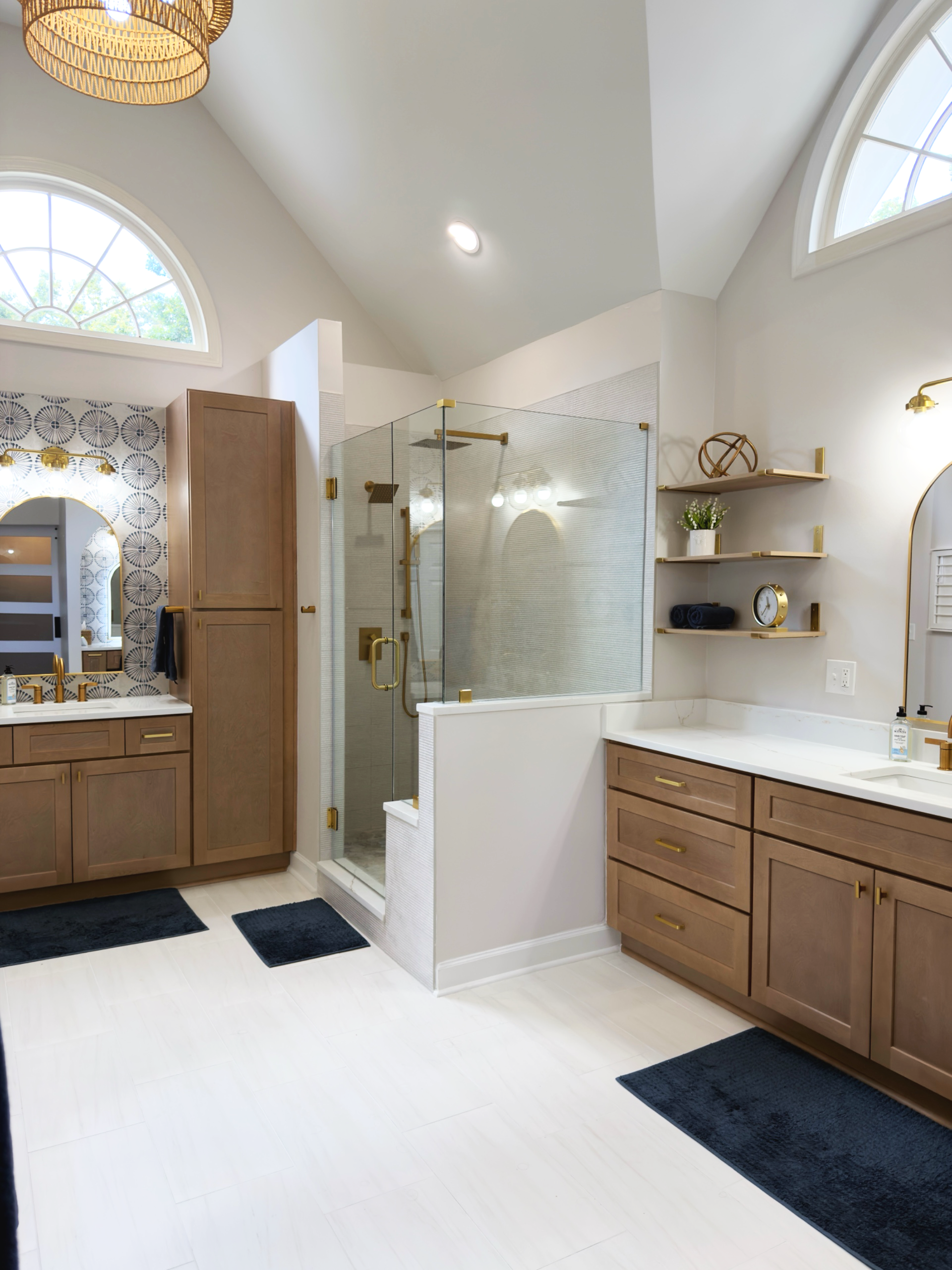 Modern bathroom with wooden cabinets, a glass shower with gold fixtures, a white vanity with a gold faucet, black rugs, decorative wall shelves with plants, towels, and a clock, and an arched window letting in natural light.