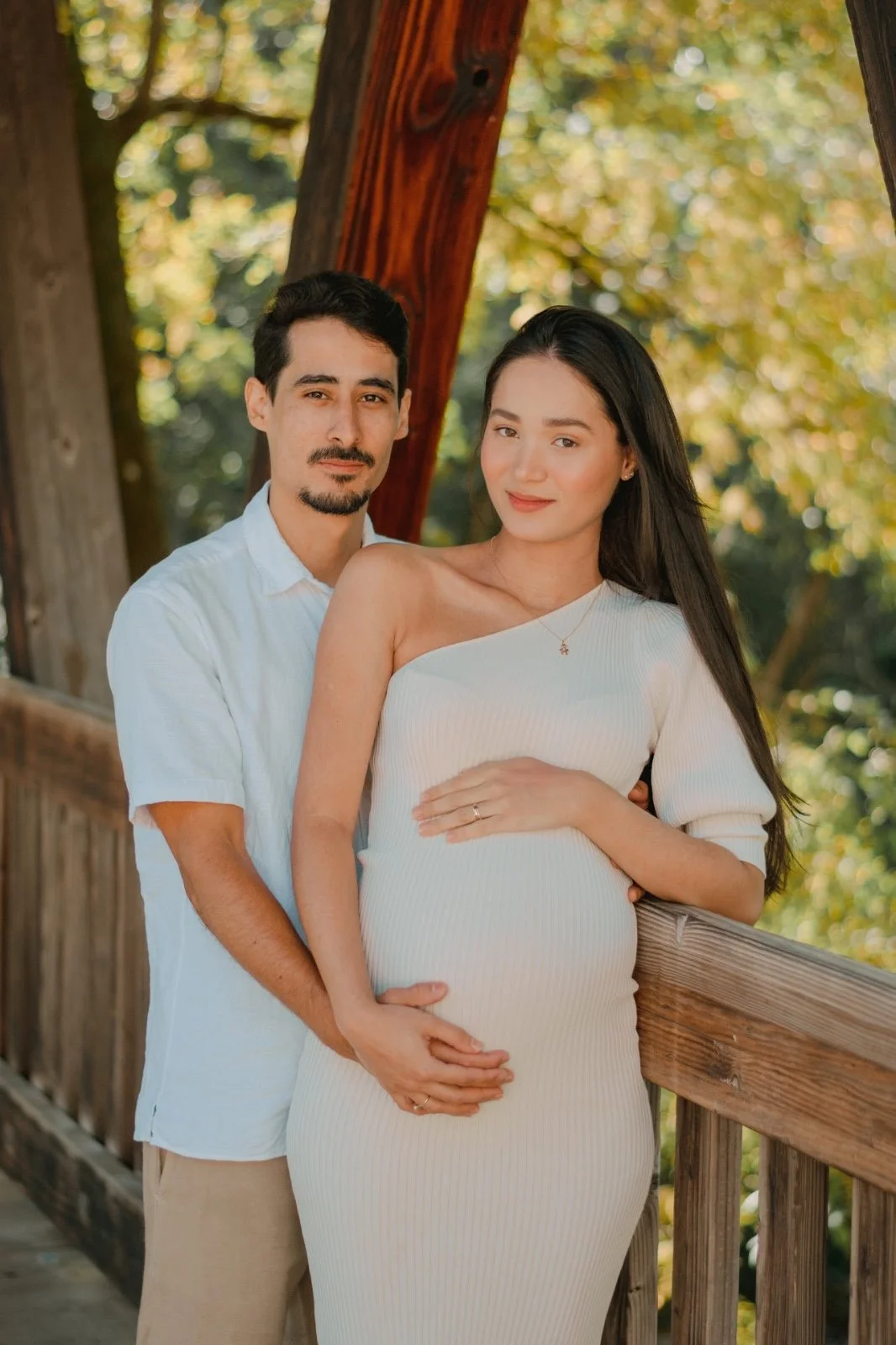 A pregnant woman and a man standing outdoors on a wooden bridge, surrounded by green trees; the woman is wearing a white dress and a necklace, and the man is wearing a white shirt and beige pants.