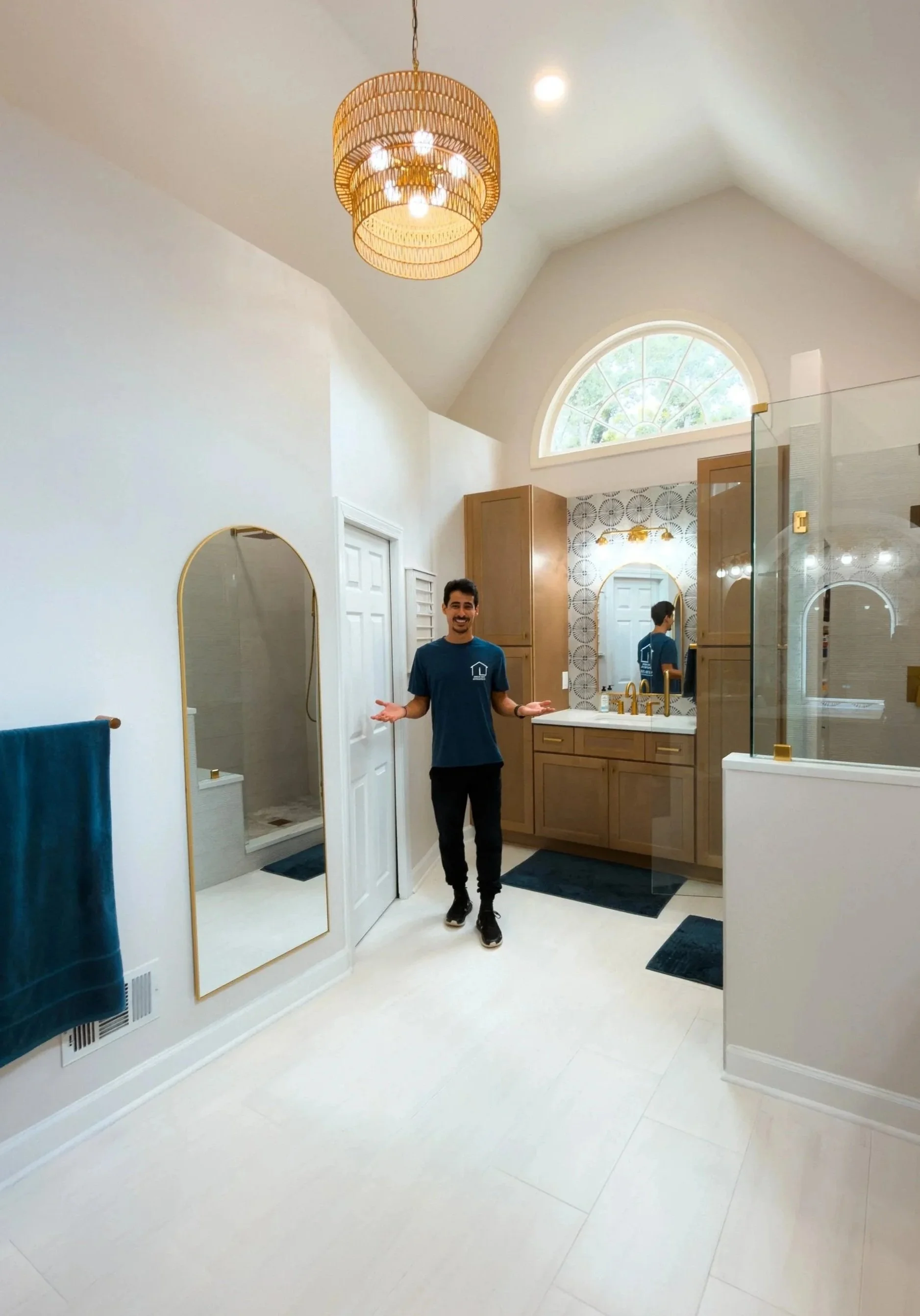 A man wearing a teal T-shirt and black pants stands smiling in a modern bathroom. The bathroom features a high, vaulted ceiling with a large, semi-circular window, a wooden vanity with a mirror and gold fixtures, a glass shower enclosure, and a decorative wall with a patterned tile. There's a blue towel hanging on a wall-mounted rack and a black bathmat on the floor.