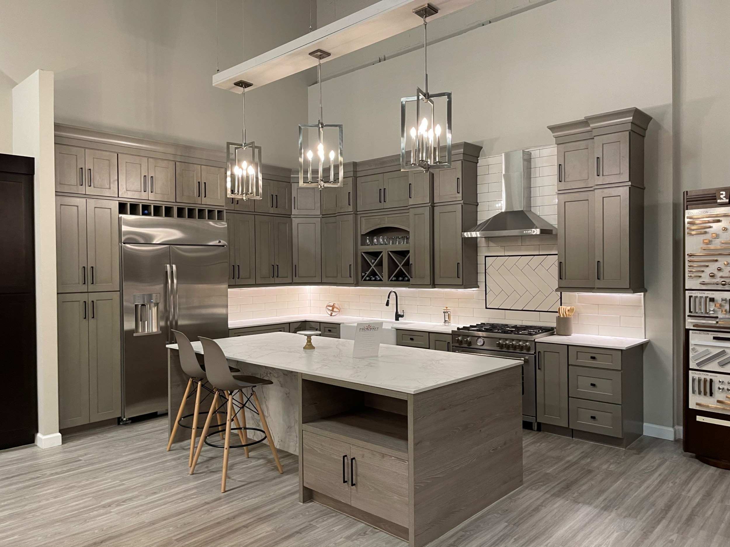 Modern kitchen with gray cabinets, stainless steel refrigerator, gas stove with stainless steel hood, and a central island with a marble countertop and two modern gray bar stools. Pendant lights hang above the island.
