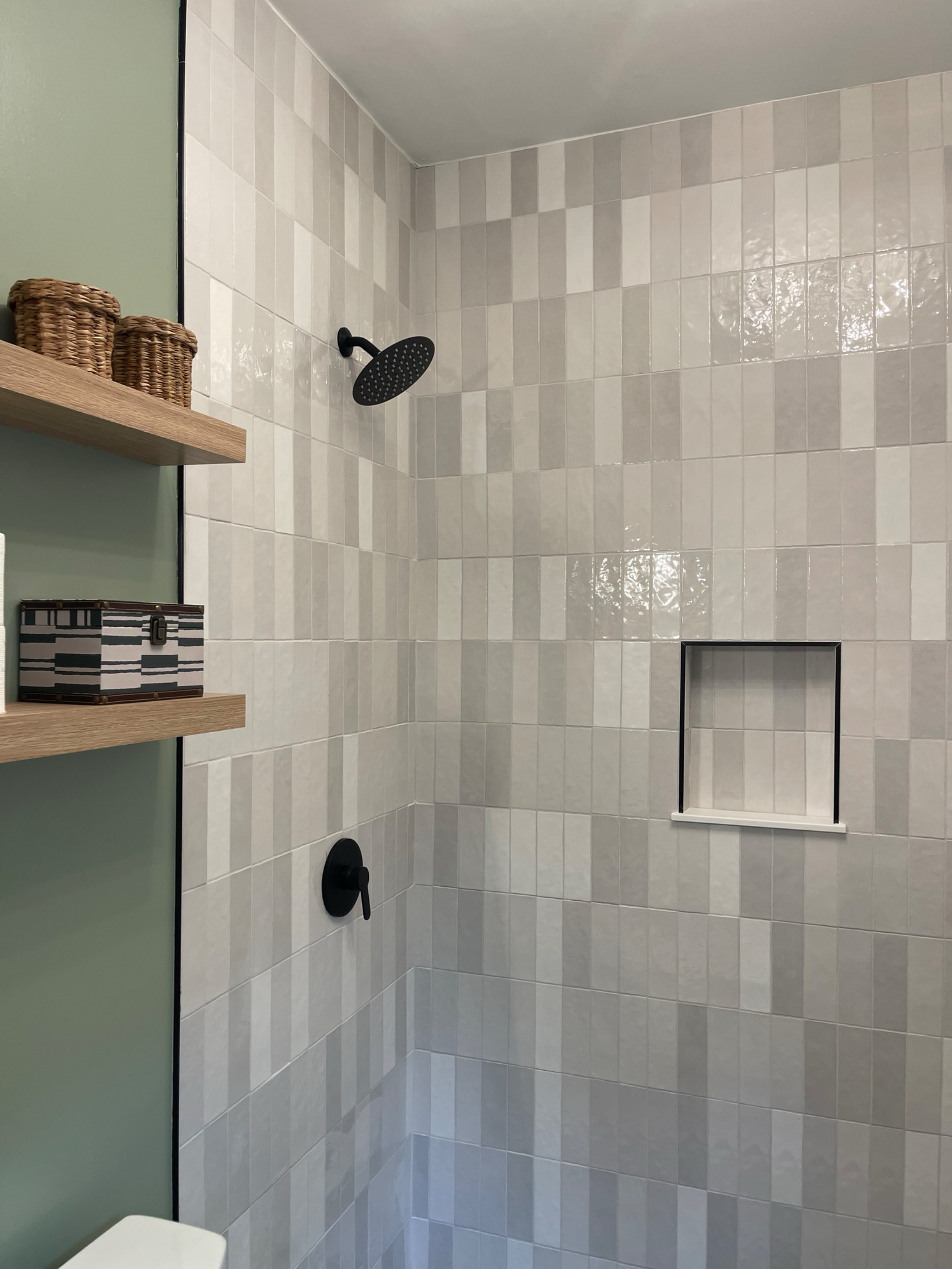 A modern shower with black fixtures, square beige and white tiles, a built-in shelf, and wooden shelves with woven baskets.