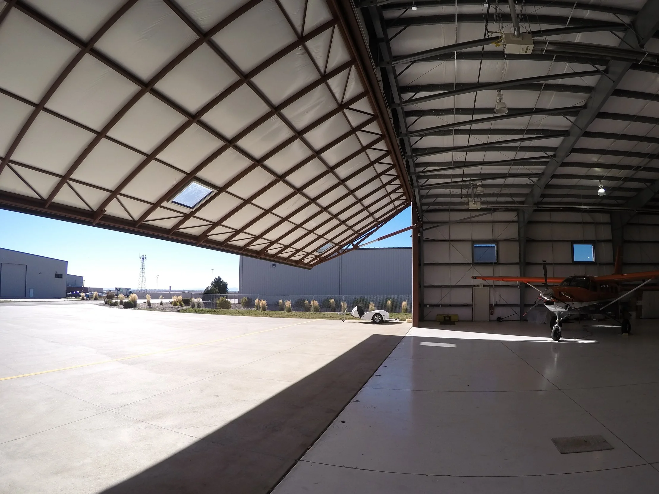 Inside a large aircraft hangar with an open door, showing a small propeller plane parked inside and an outdoor concrete tarmac with a small white car and industrial buildings in the background.