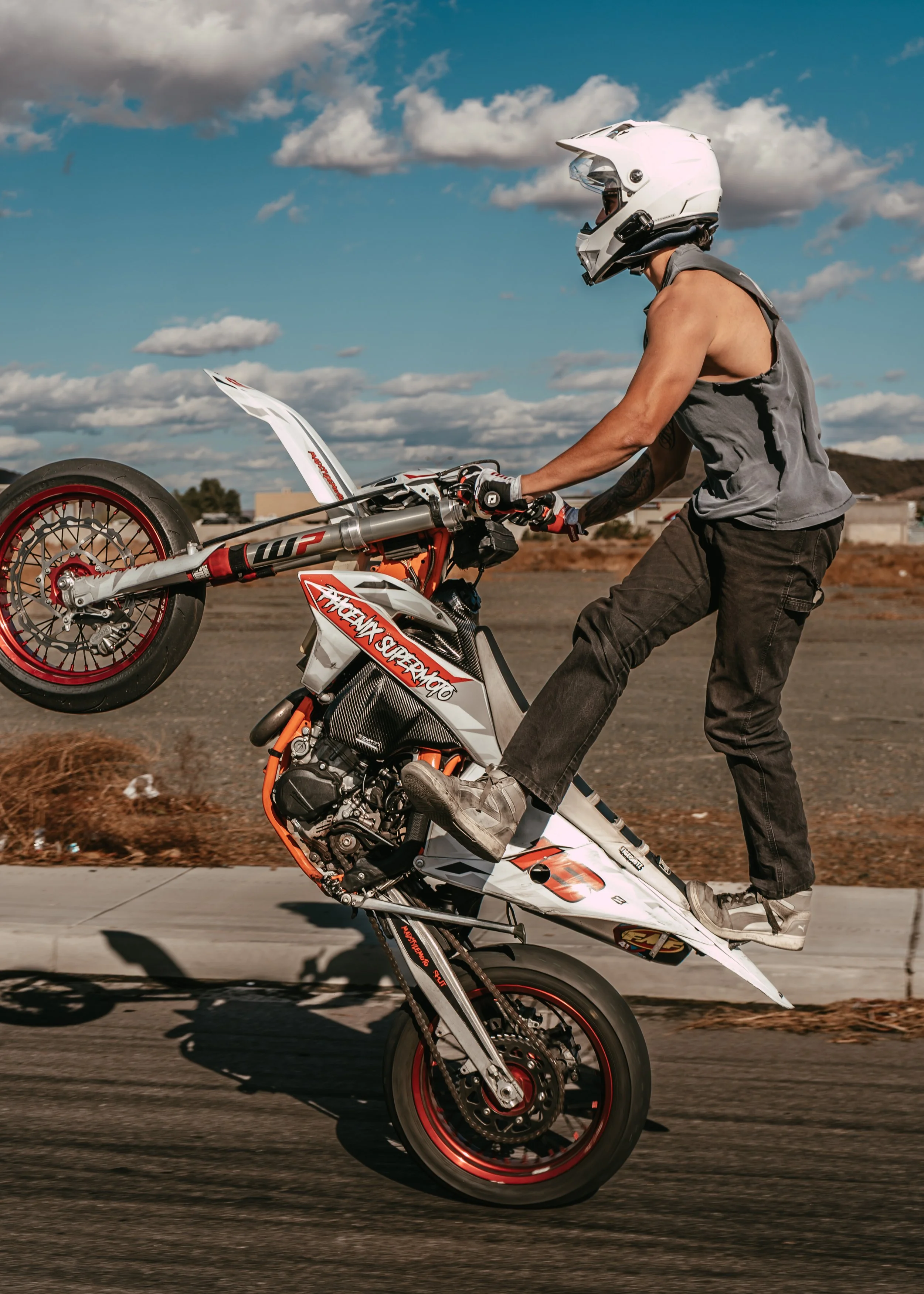 A person performing a wheelie on a custom motocross-style dirt bike on a paved road, wearing a white helmet, gray sleeveless shirt, black pants, and gloves.