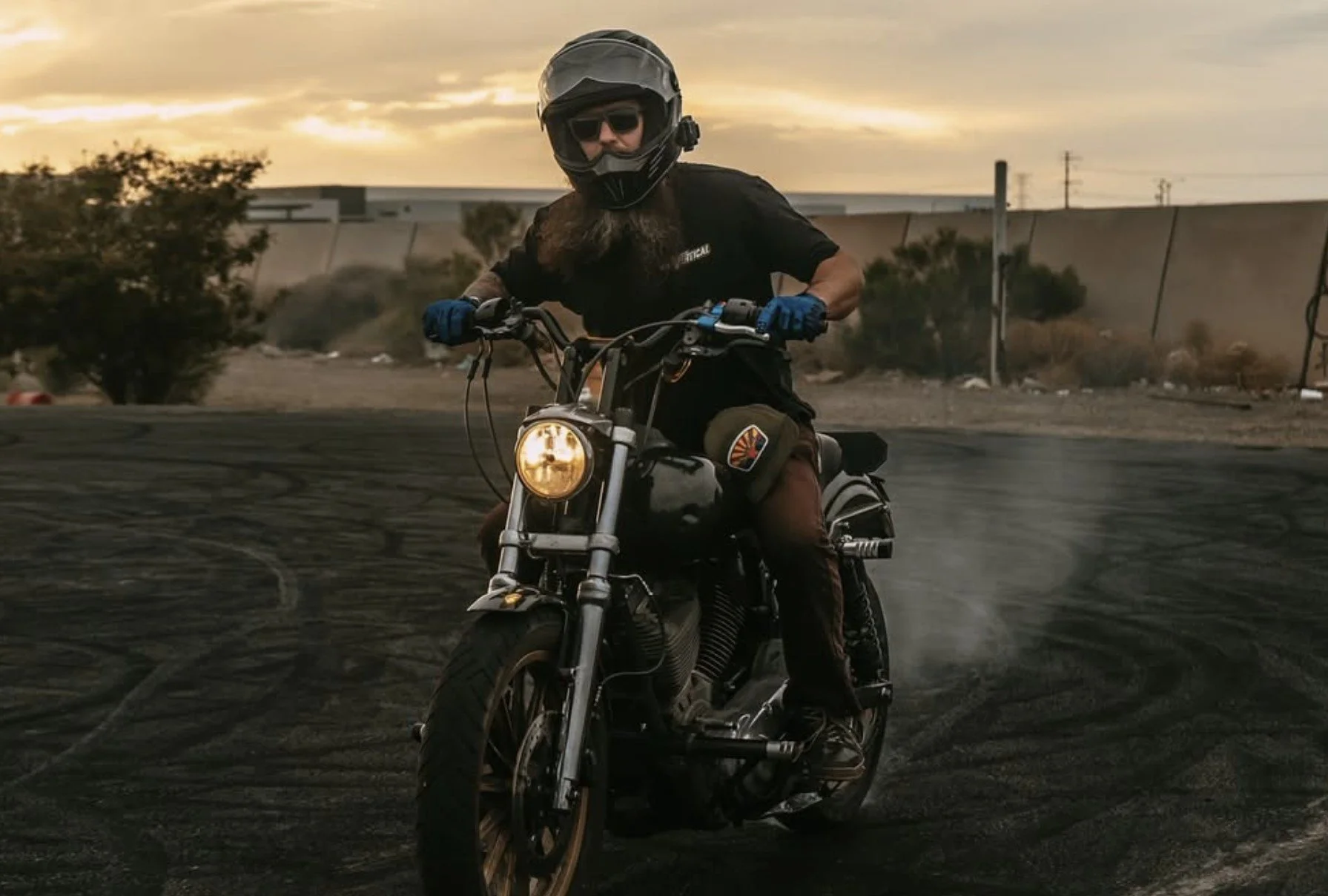 A man riding a motorcycle on a dirt road during sunset, wearing a helmet, sunglasses, black t-shirt, and gloves.