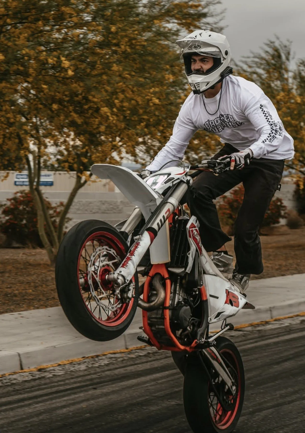 Person wearing a white helmet and shirt performing a wheelie on a motorcycle on a paved road with trees and bushes in the background.