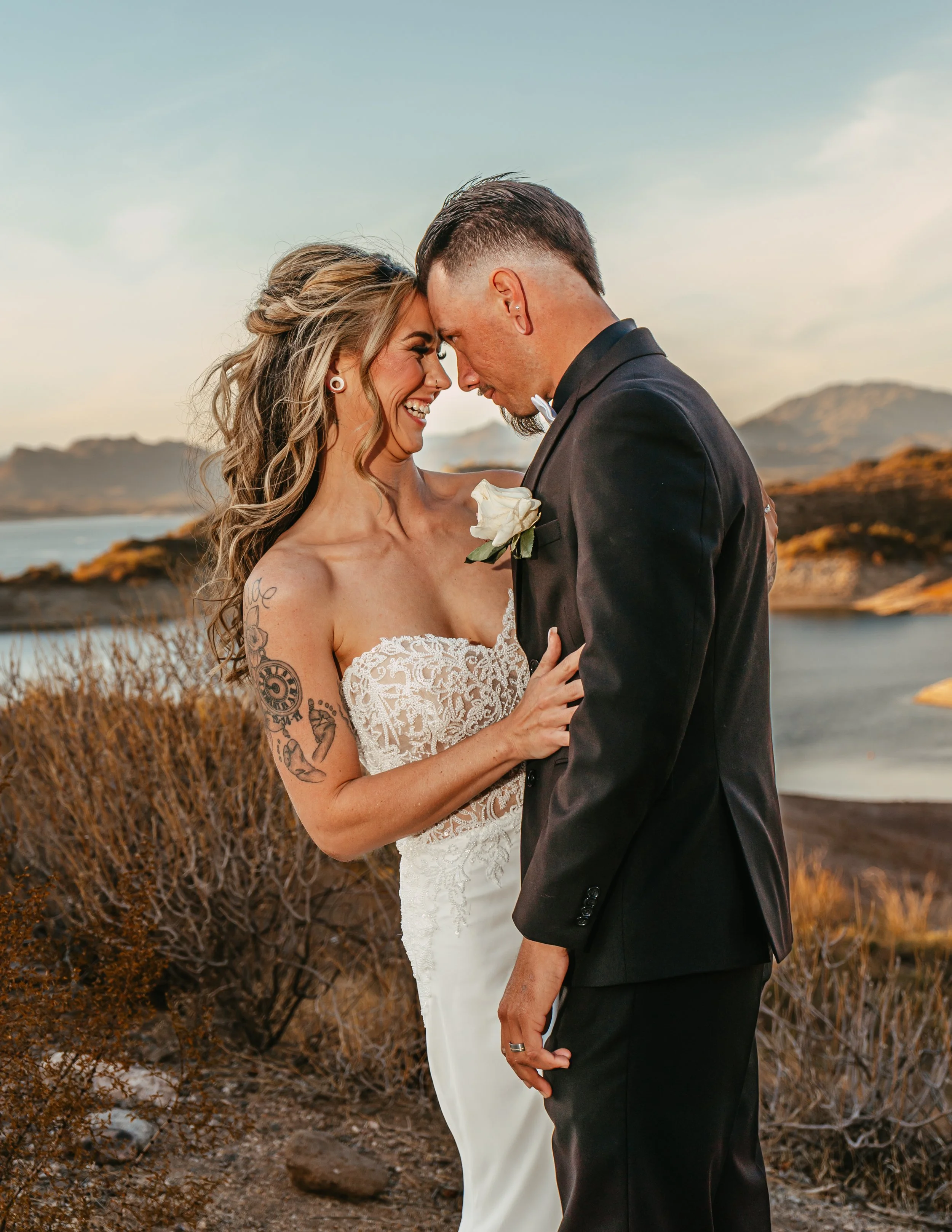 A newlywed couple shares a close moment outdoors near a lake at sunset, with mountains in the background. The bride has tattoos on her arm and is wearing a strapless, lace wedding dress. The groom is in a black tuxedo with a white boutonniere. They are smiling and touching foreheads.