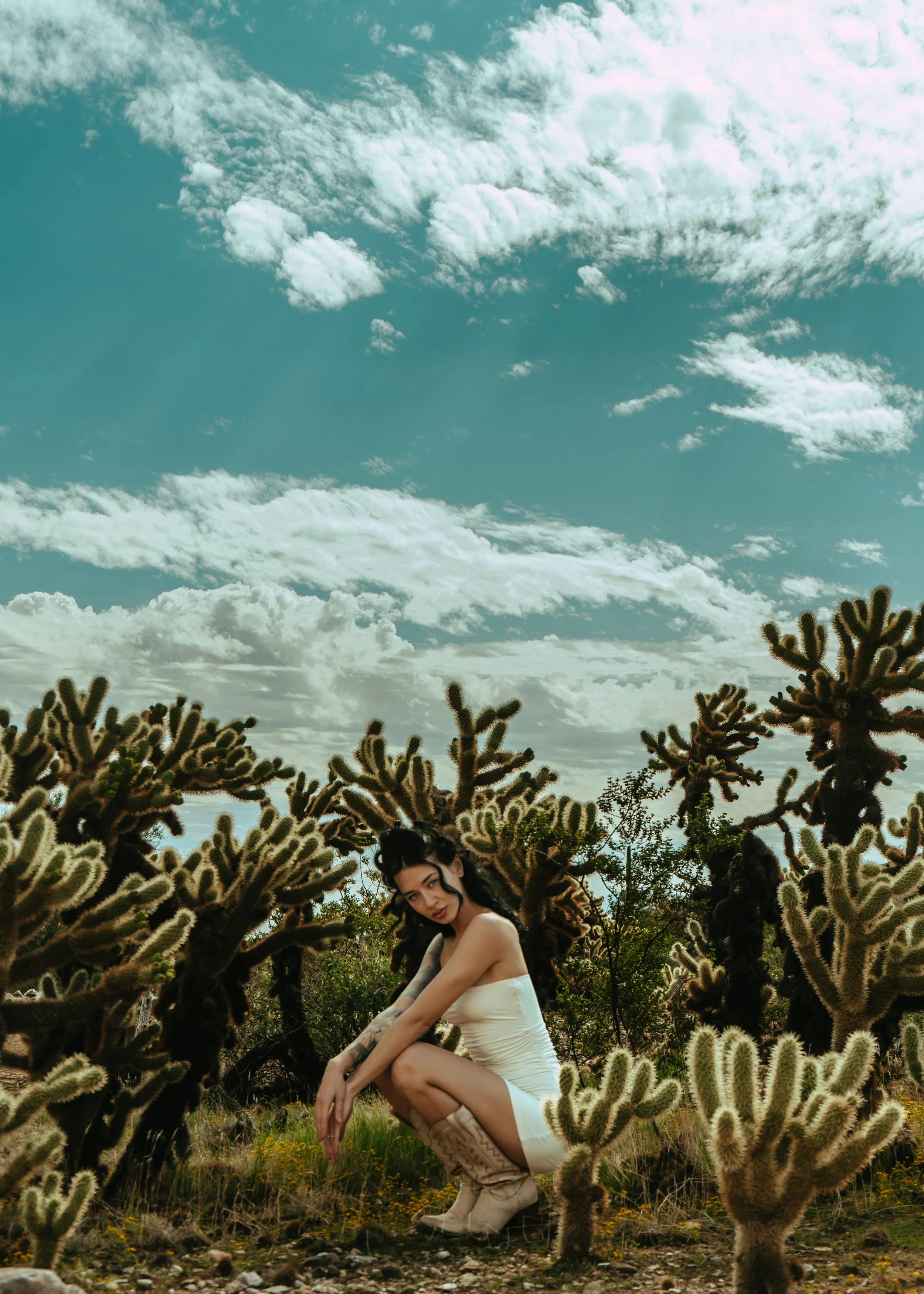 A woman in a white dress crouching among cactus plants outdoors under a partly cloudy sky.