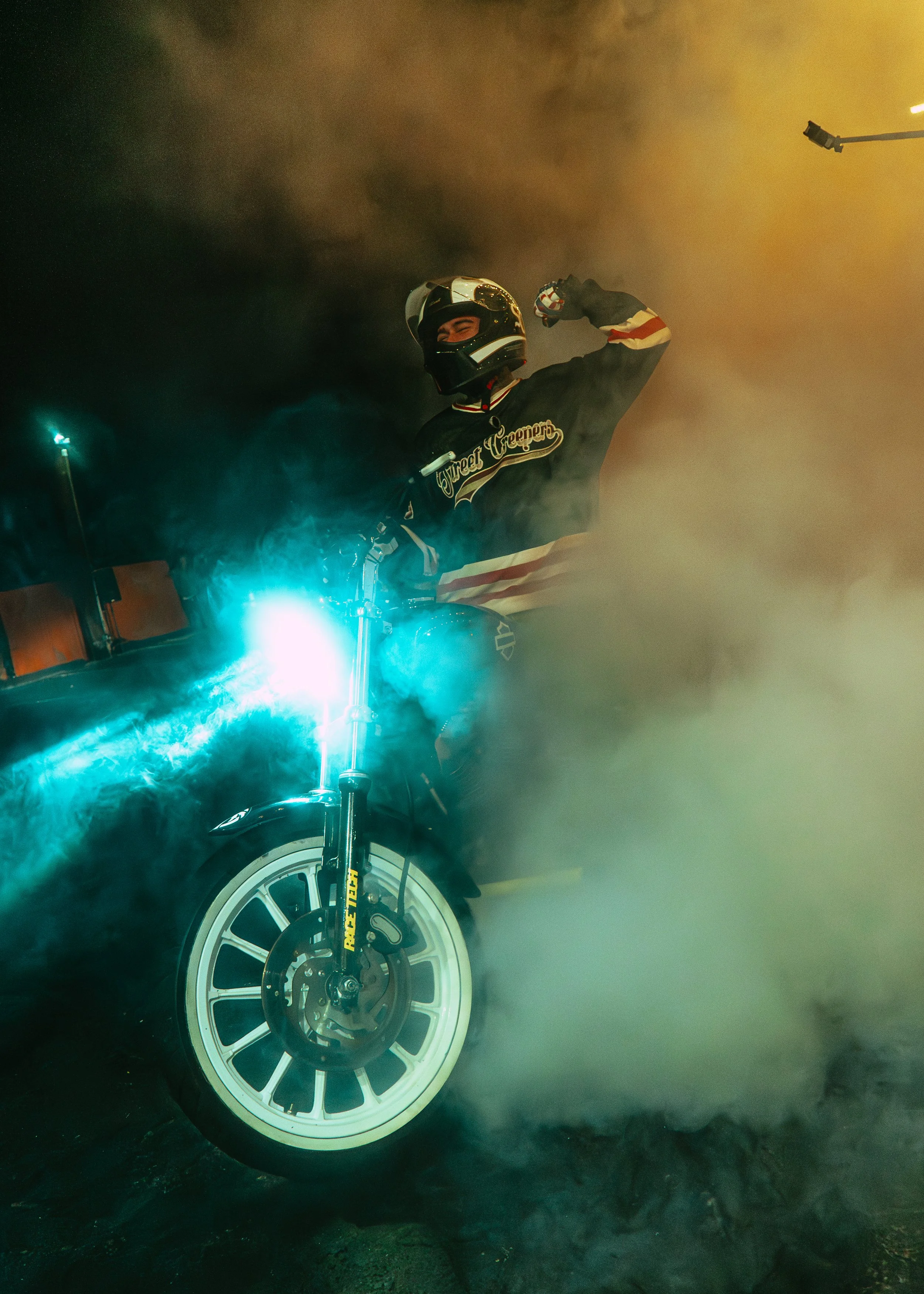 A person wearing a helmet and jacket riding a motorcycle through smoke, with blue lights reflecting off the motorcycle.