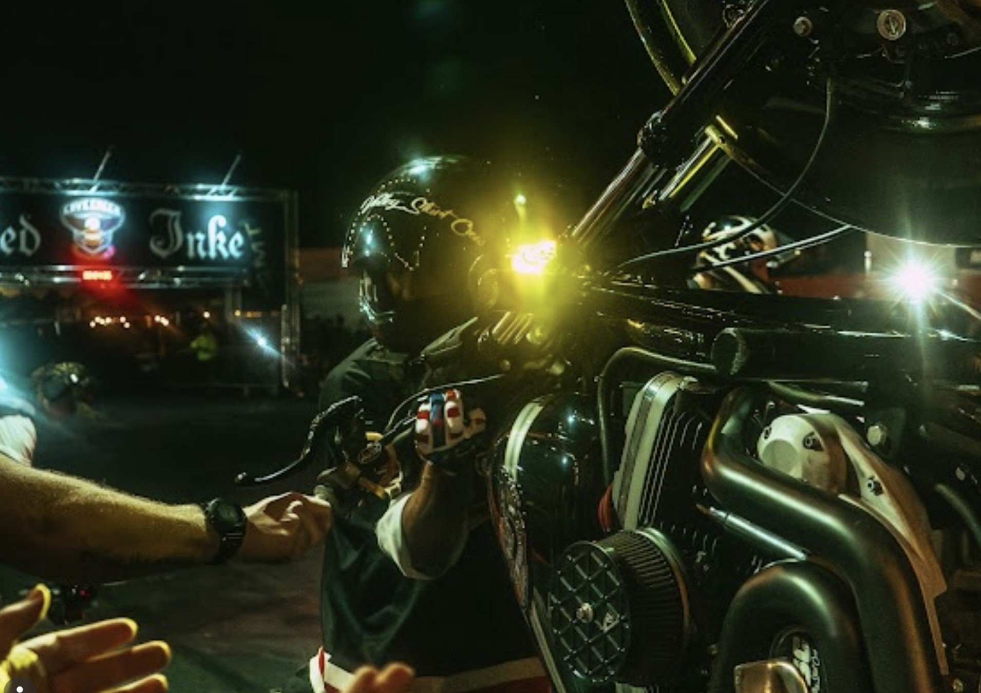 A motorcycle racer wearing a black helmet with sponsorship logos, preparing on the starting grid at night, with team members surrounding, and the 