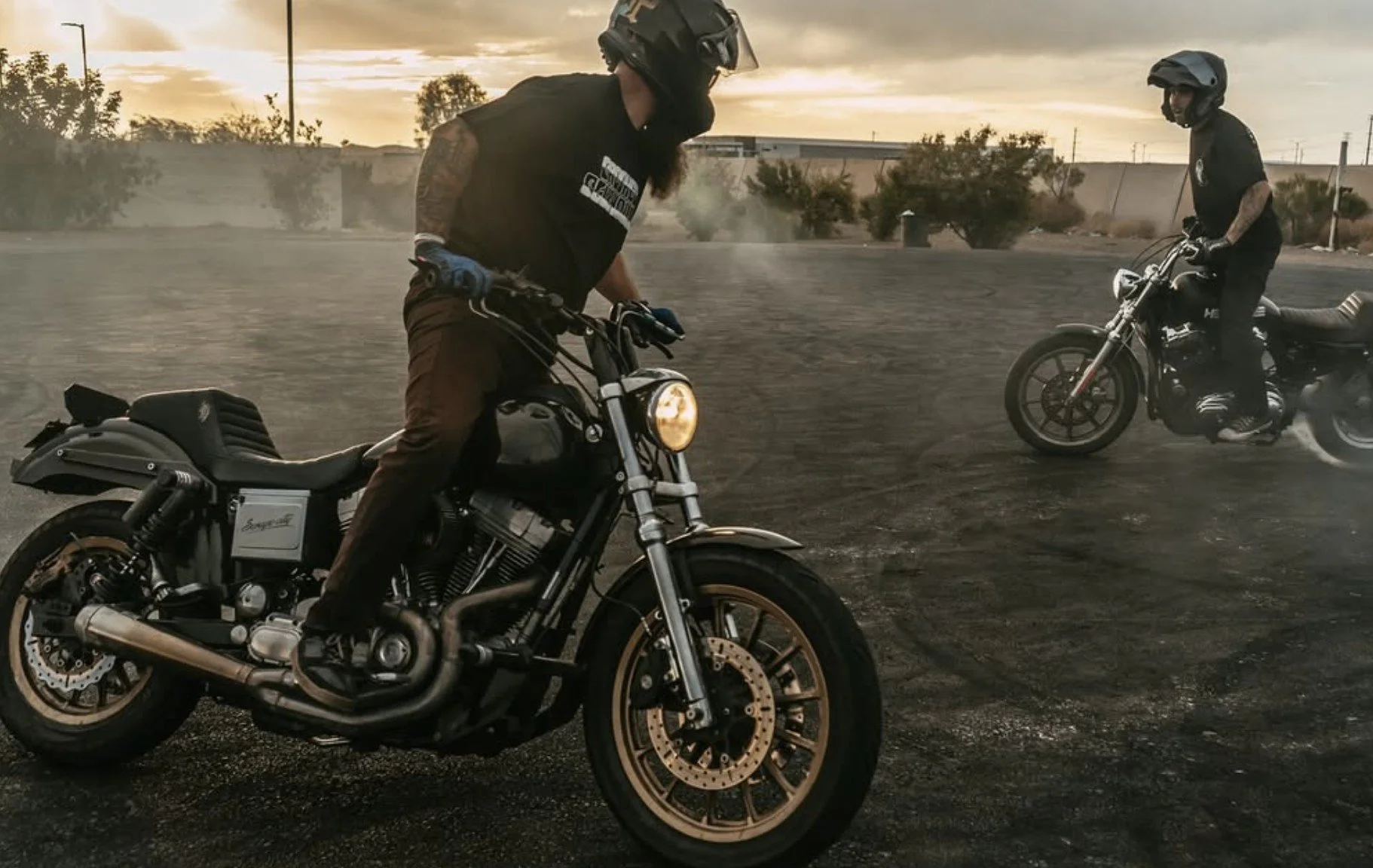 Two motorcyclists riding on a dirt lot during sunset, with one rider in the foreground and another in the background, both wearing helmets and black clothing.