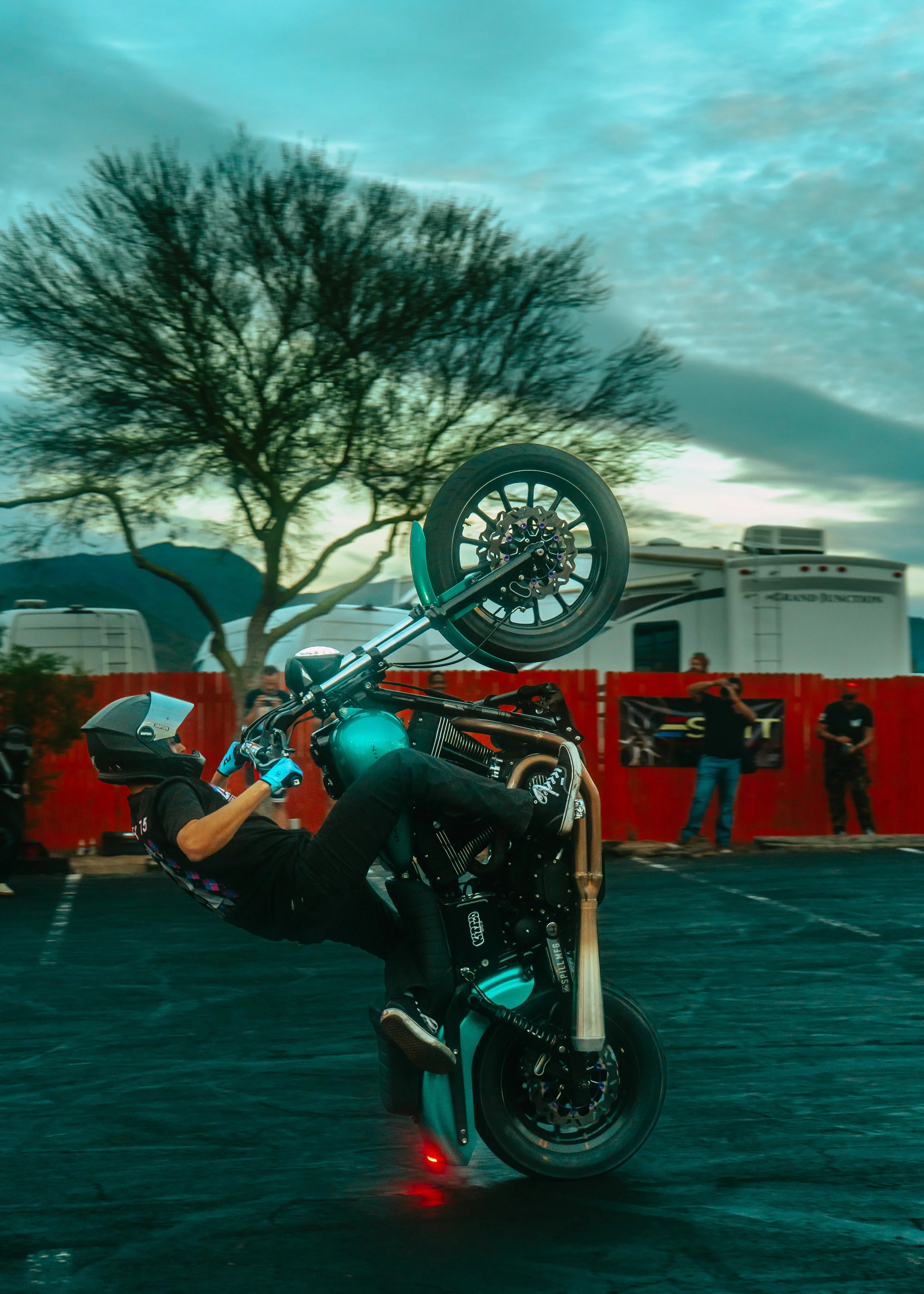 Motorcyclist performing a wheelie stunt on a motorcycle in an outdoor setting with trees and a red fence in the background.