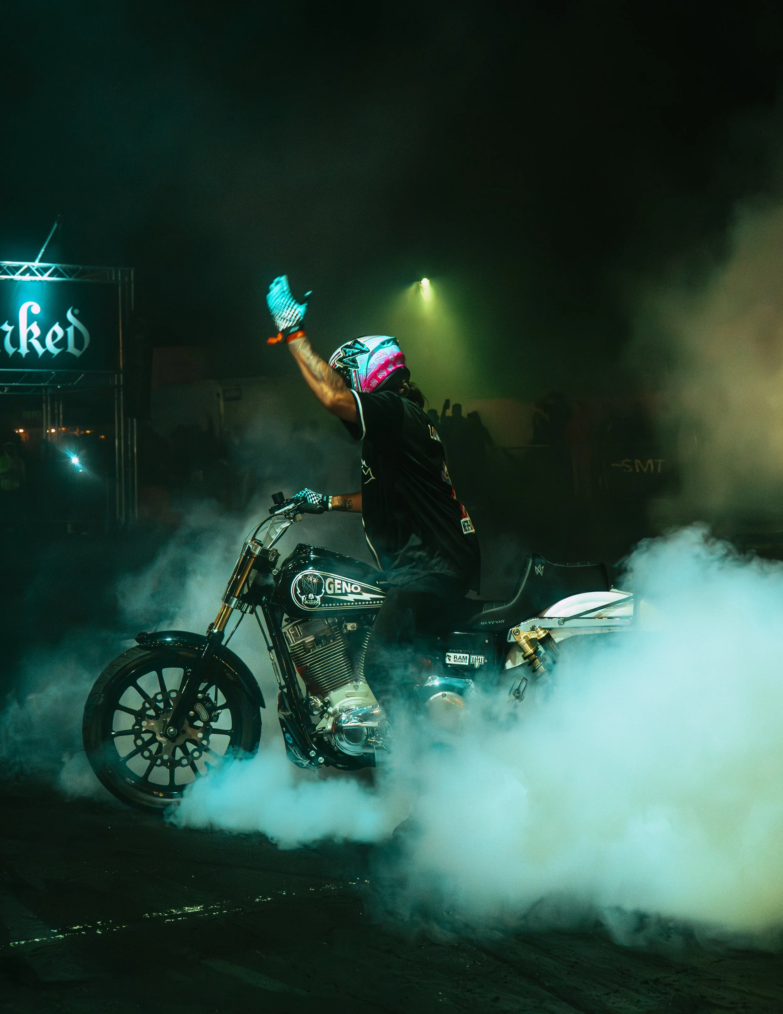 Person wearing a helmet and black outfit performing a burnout on a black motorcycle with smoke billowing around, at night with green and yellow lighting.