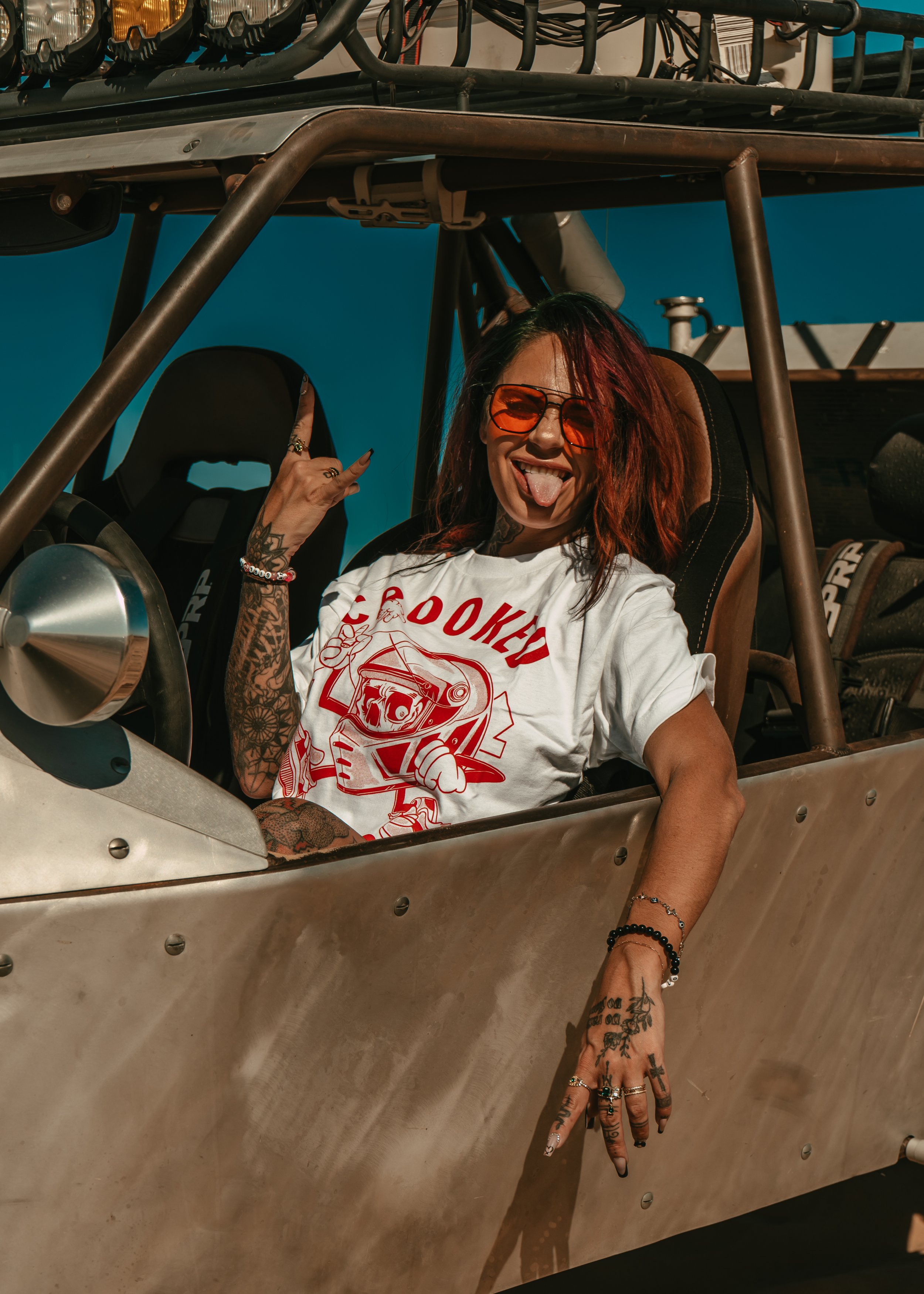 A woman with tattoos, red hair, and sunglasses sitting in a race car, making a rock sign and sticking out her tongue.